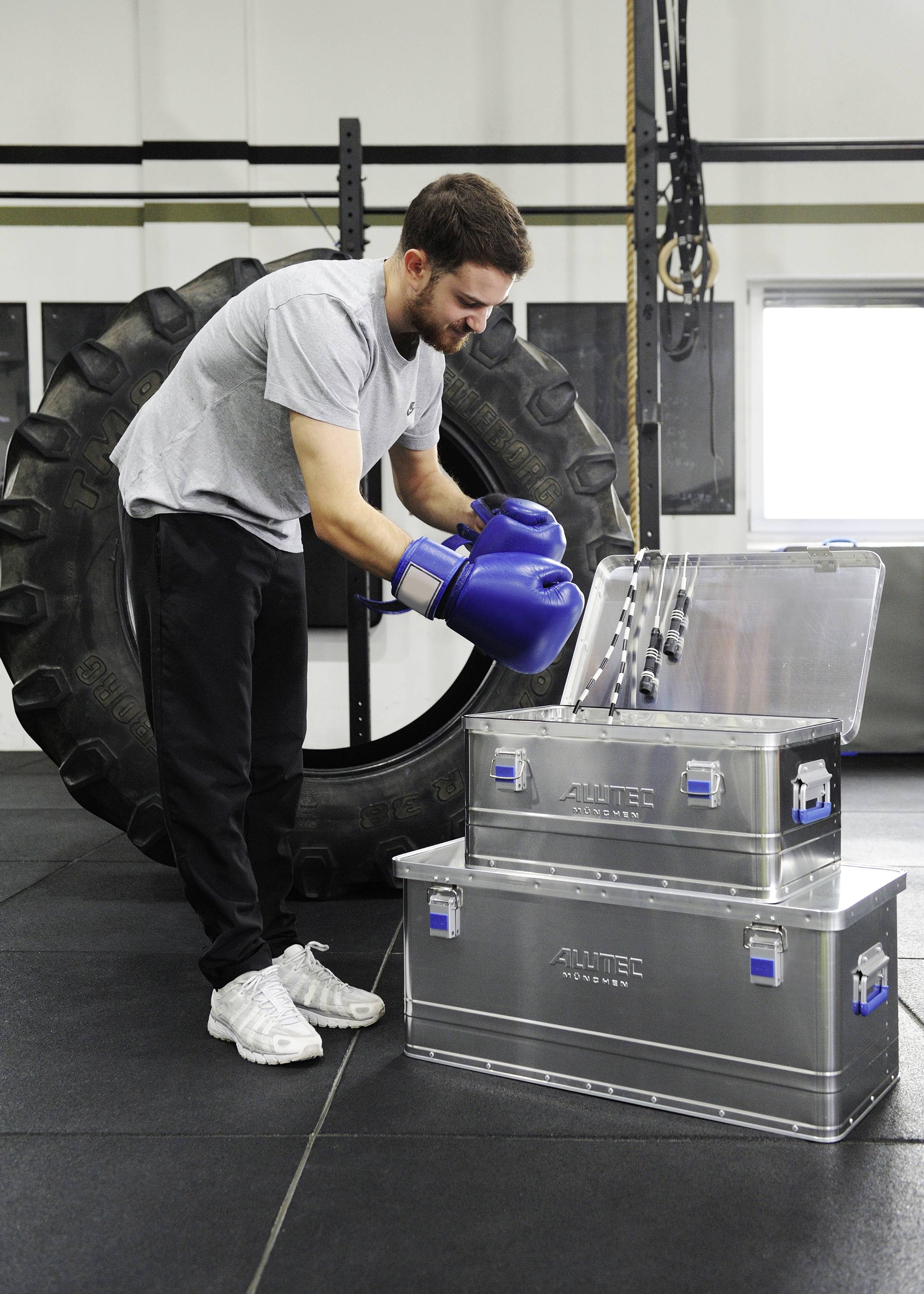 Ein Mann in Sportkleidung und Boxhandschuhen steht in einem Fitnessstudio vor großen Metallkisten und einem Traktorreifen. Er öffnet eine der Kisten.