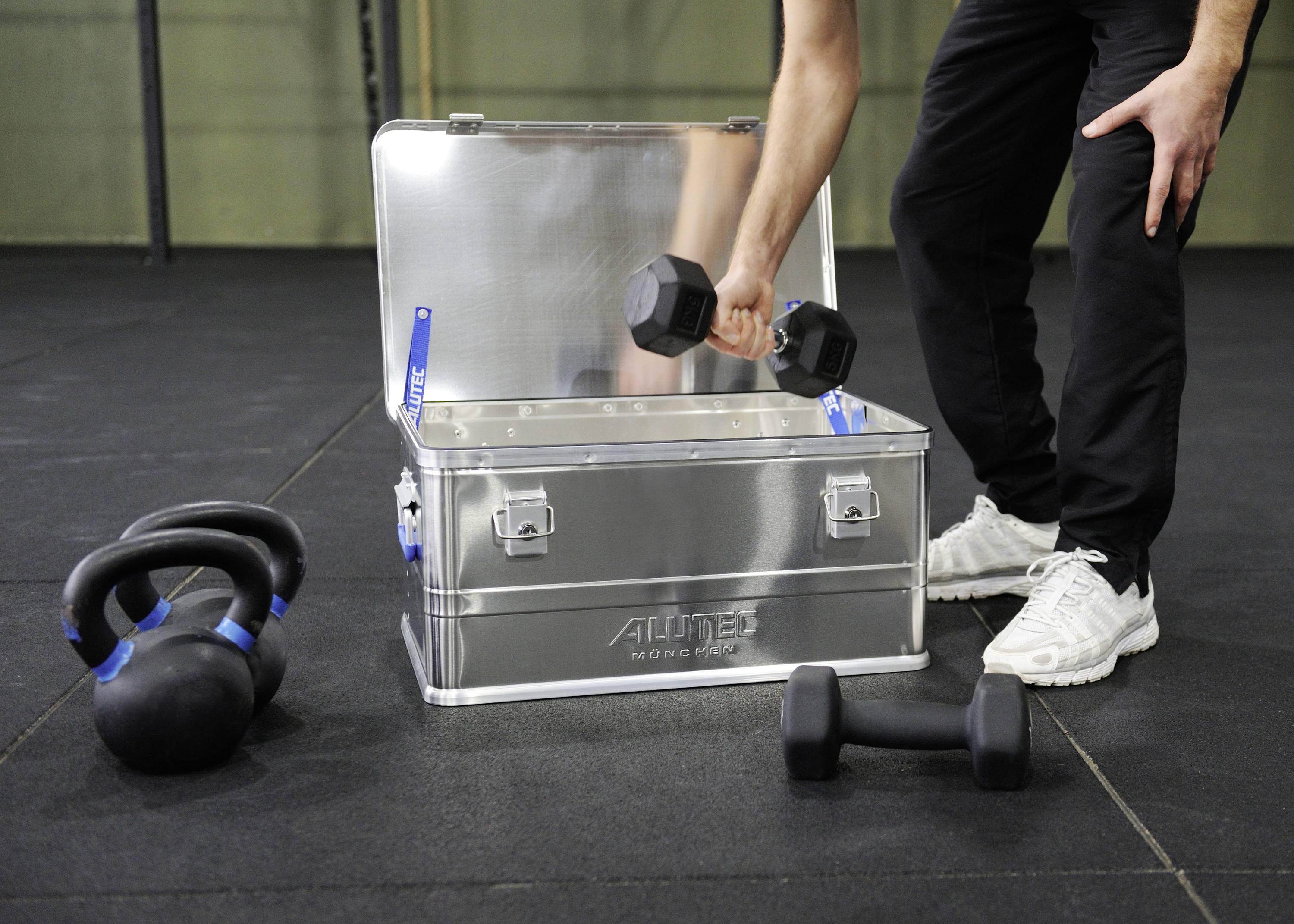 Eine Person hebt eine Hantel aus einer offenen Metallkiste auf einem Fitnessstudio-Boden an. Neben der Kiste liegen Kettlebells.