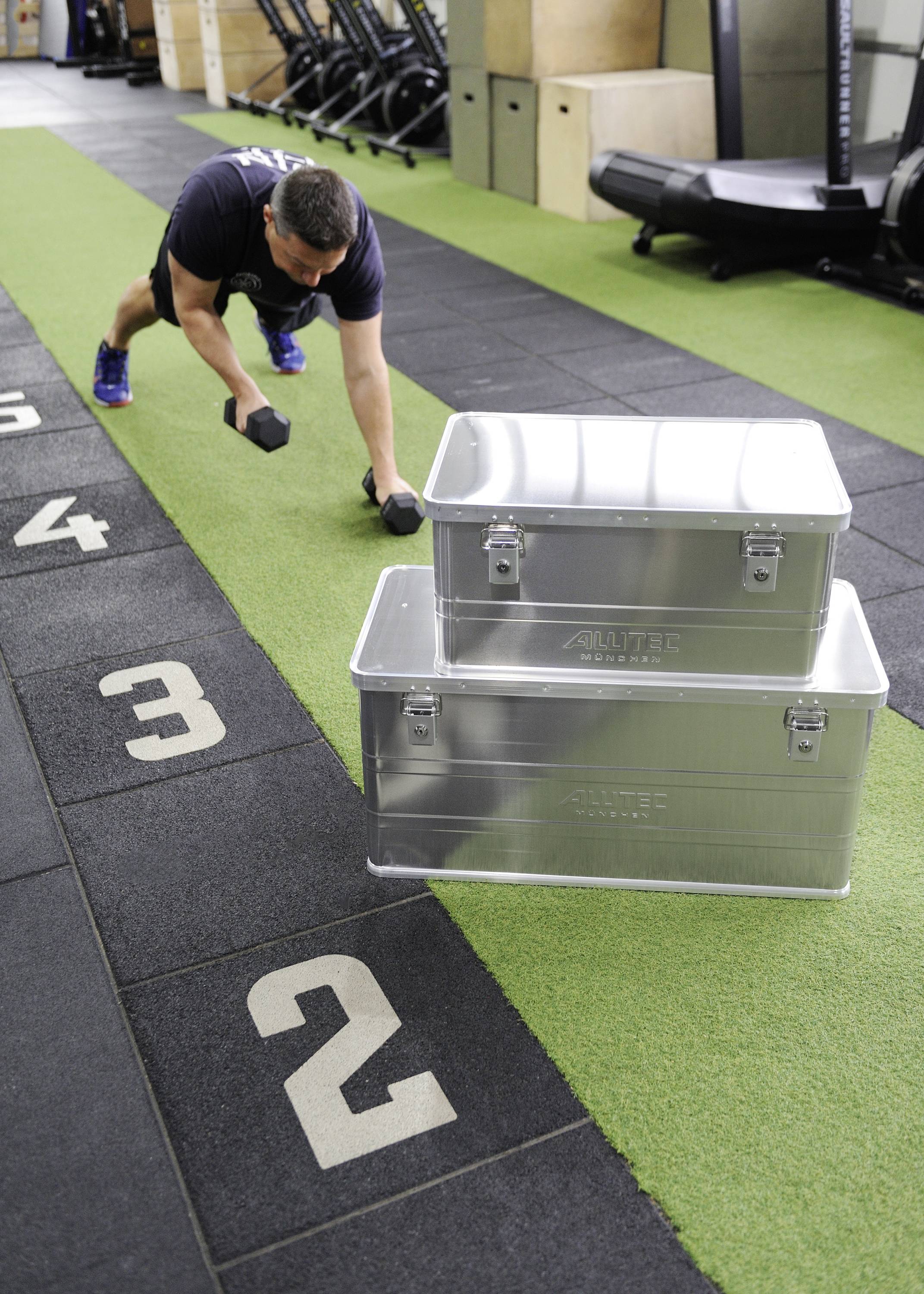 Ein Mann macht Liegestütze mit Hanteln neben gestapelten Metallboxen in einem Fitnessstudio. Boden mit nummerierten Markierungen.