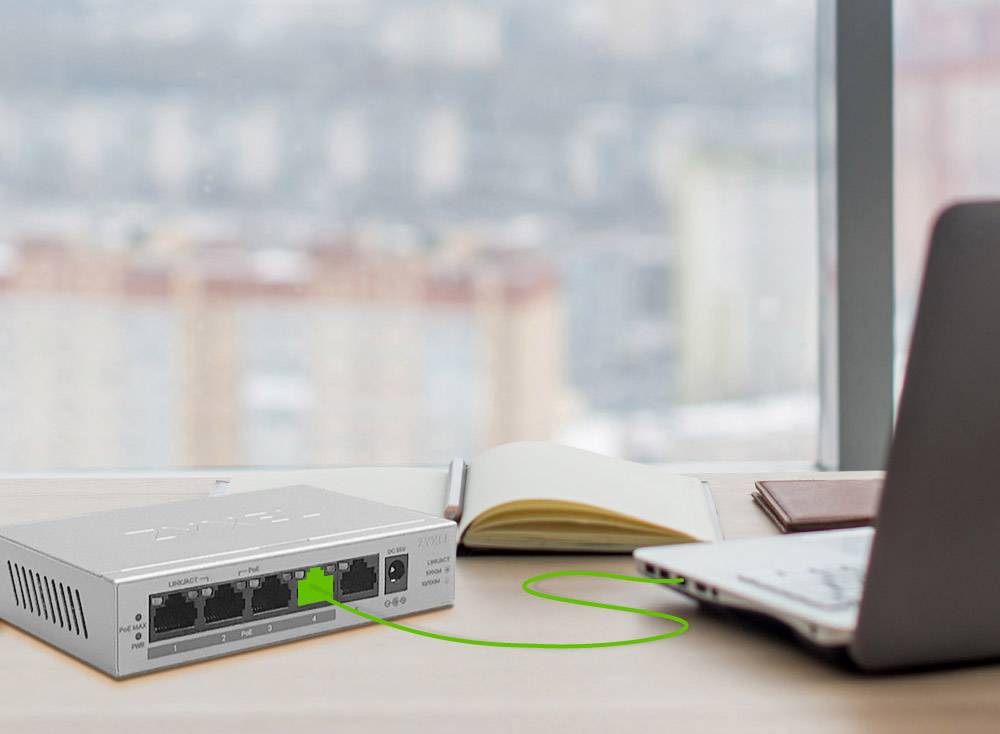 Ein Laptop auf einem Schreibtisch ist mit einem Netzwerk-Switch verbunden. Im Hintergrund ist eine unscharfe städtische Landschaft zu sehen.