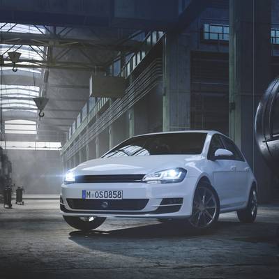 Ein weißes Auto steht in einer beleuchteten Lagerhalle. Es ist von vorne rechts sichtbar. Fenster und Decke haben Metallstrukturen.