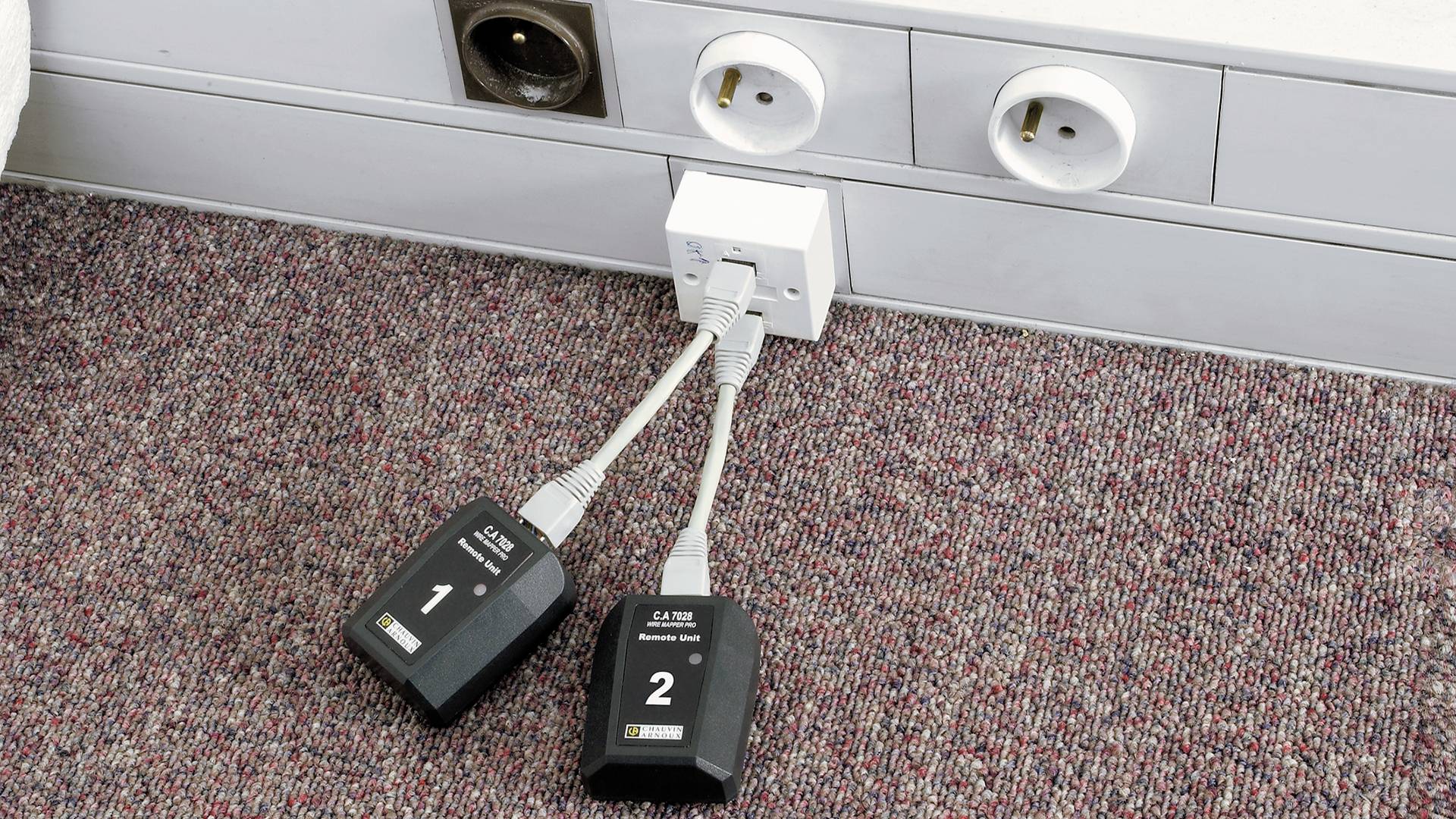 Zwei elektronische Geräte mit Beschriftungen '1' und '2' sind via Kabel mit einer Wandsteckdose verbunden, die auf einem Teppichboden montiert ist.