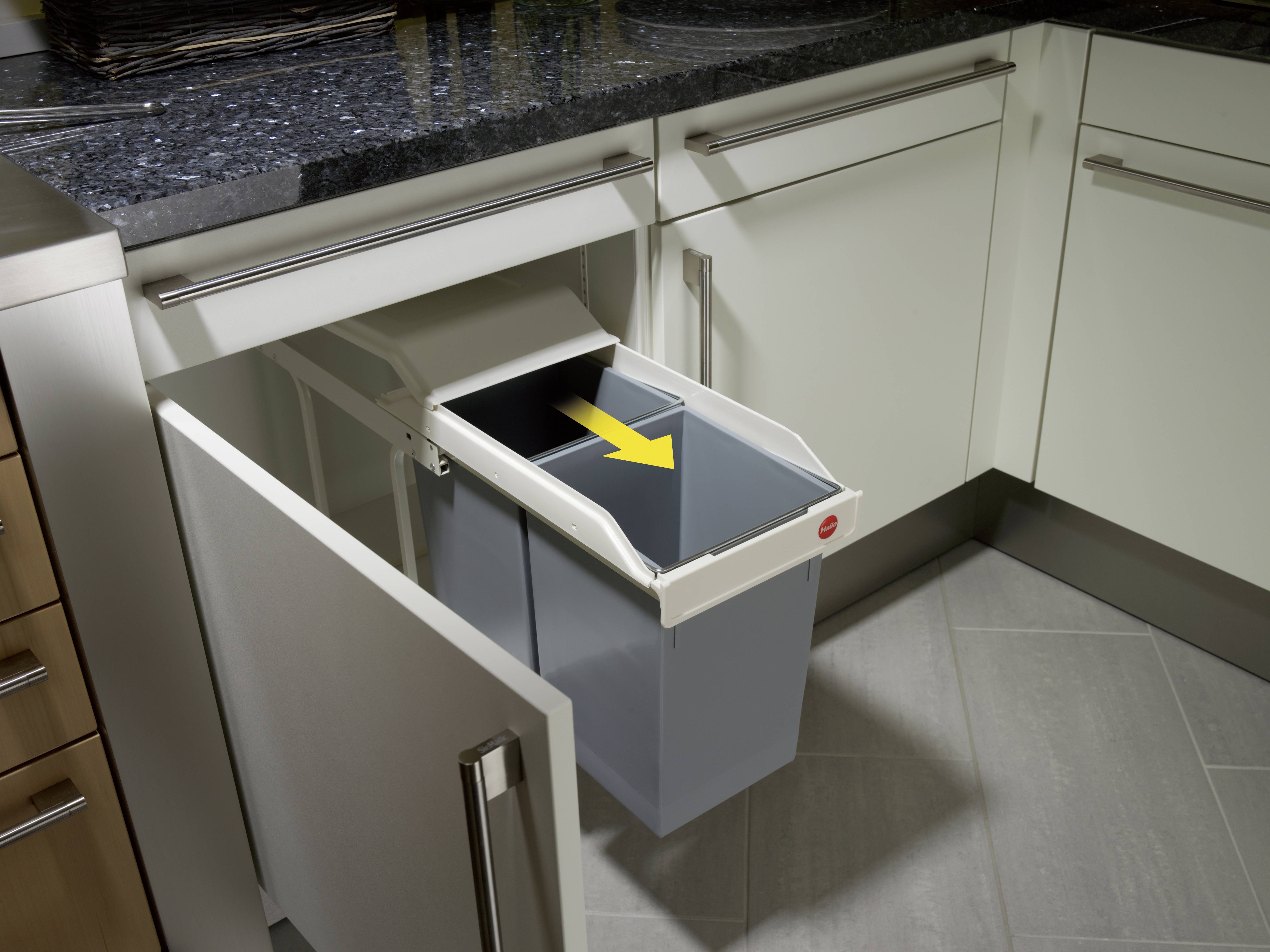 Ein geöffneter Unterschrank in einer Küche zeigt einen herausziehbaren Müllbehälter mit einem gelben Pfeil, der den Schließmechanismus anzeigt.