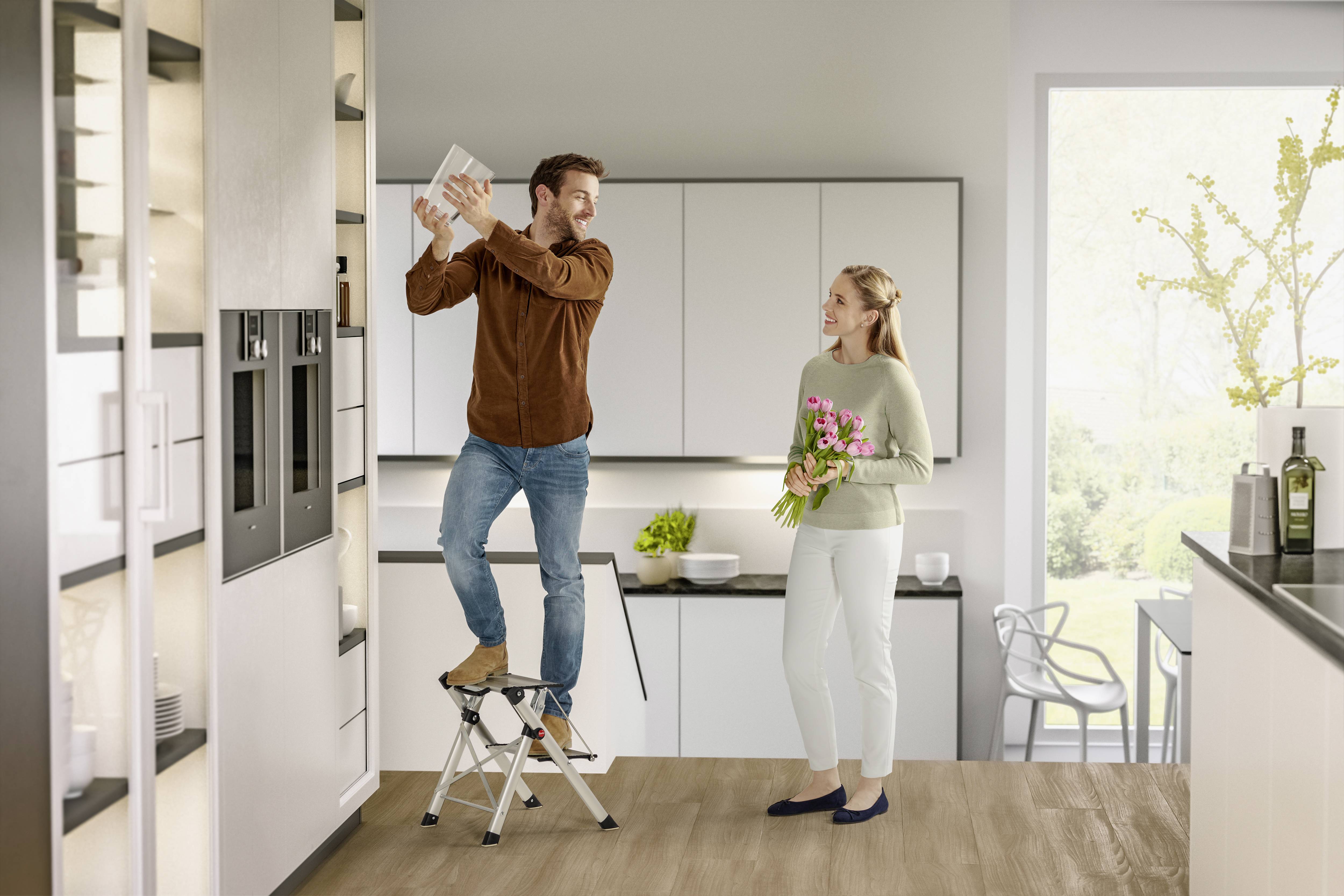 Ein Mann steht auf einer Trittleiter in einer modernen Küche und stellt etwas in den Schrank, während eine Frau mit Blumen zusieht.