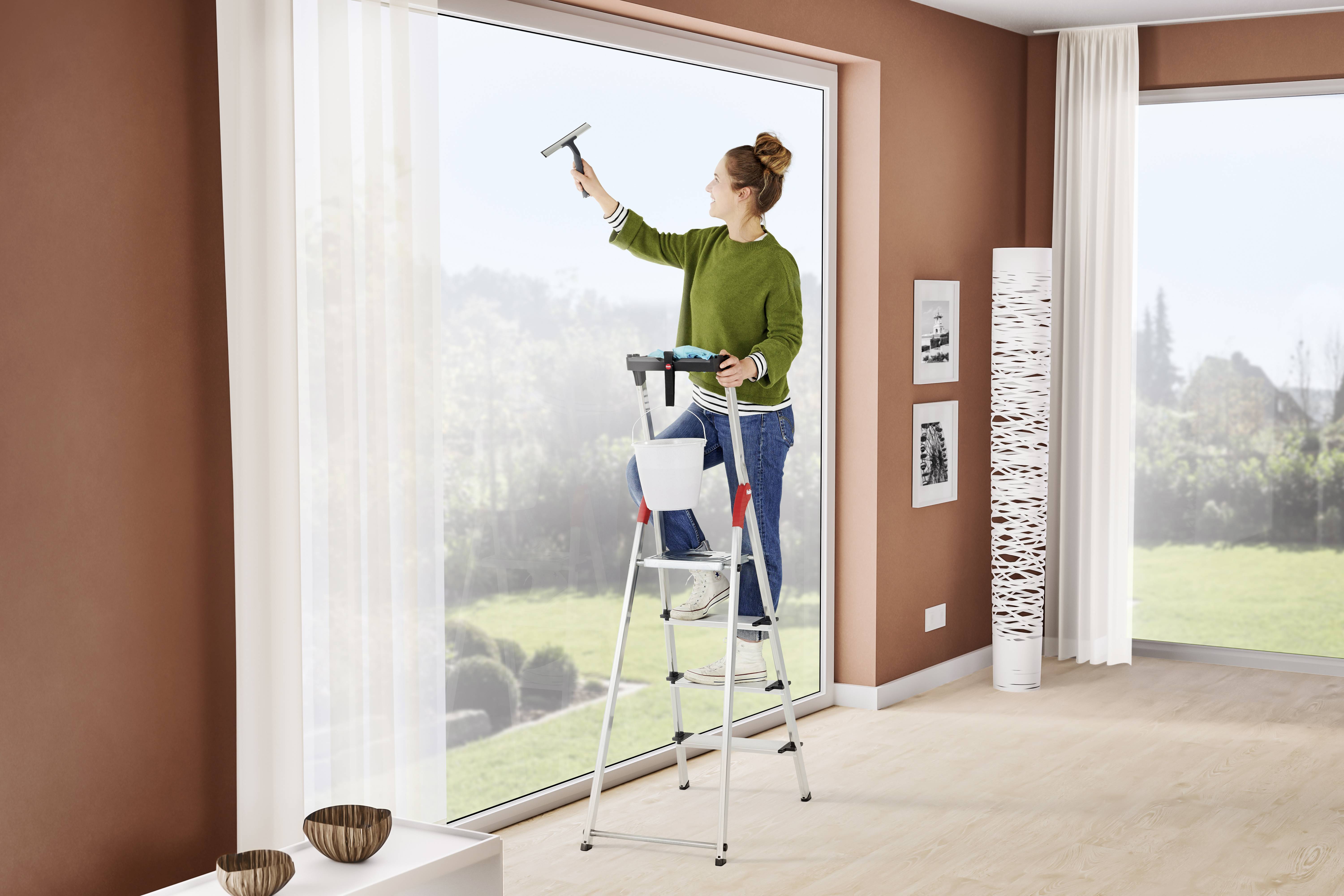 Eine Frau in grünem Pullover steht auf einer Leiter und putzt ein großes Fenster in einem hellen Raum mit Holzfußboden und Dekorationen.