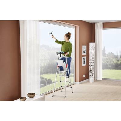 Person reinigt ein großes Fenster von innen mit einem Fensterwischer. Sie steht auf einer Leiter in einem hellen, modern eingerichteten Raum.