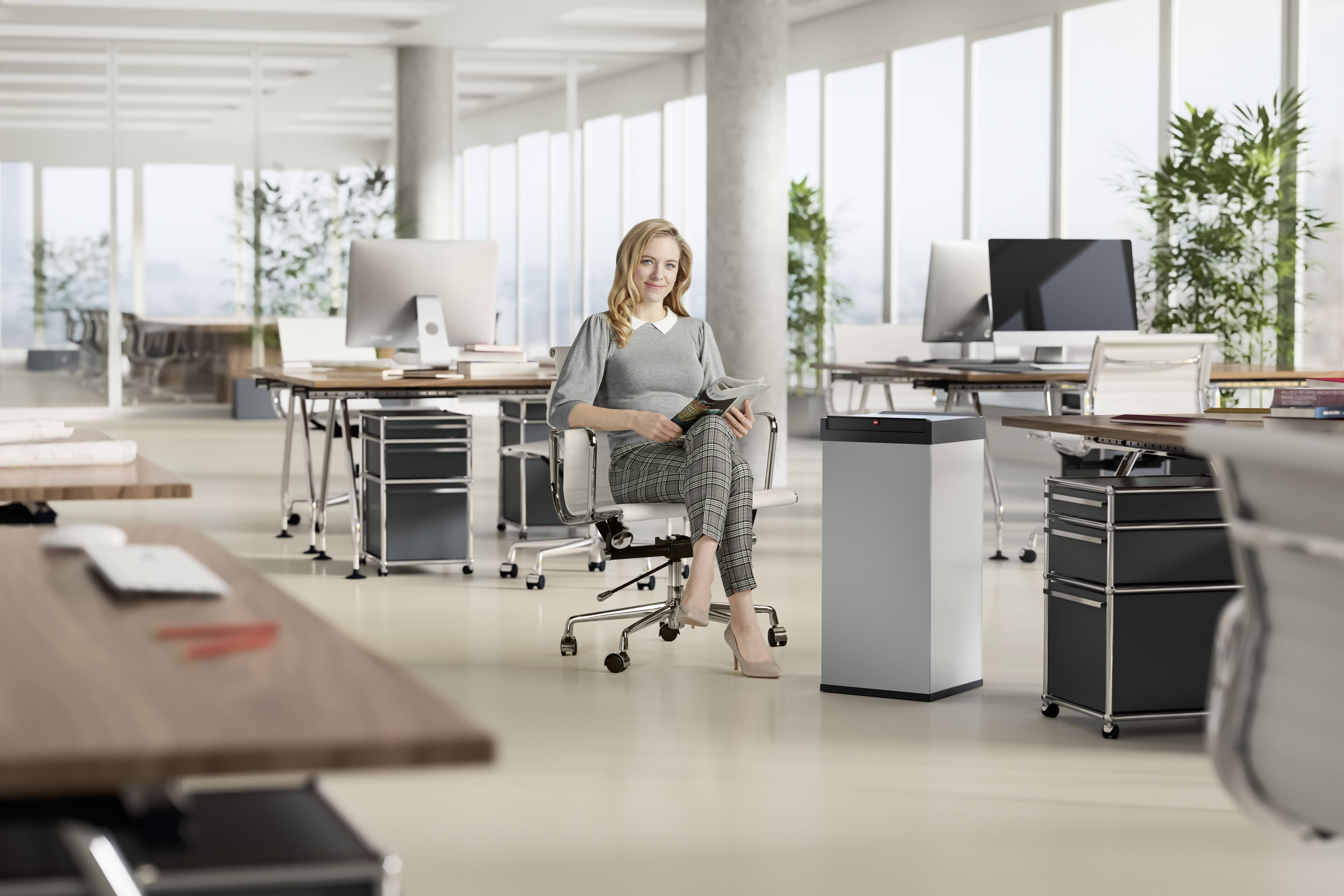 Eine Person sitzt in einem modernen Büro mit mehreren Schreibtischen und Computern, während sie ein Heft hält. Pflanzen stehen im Hintergrund.