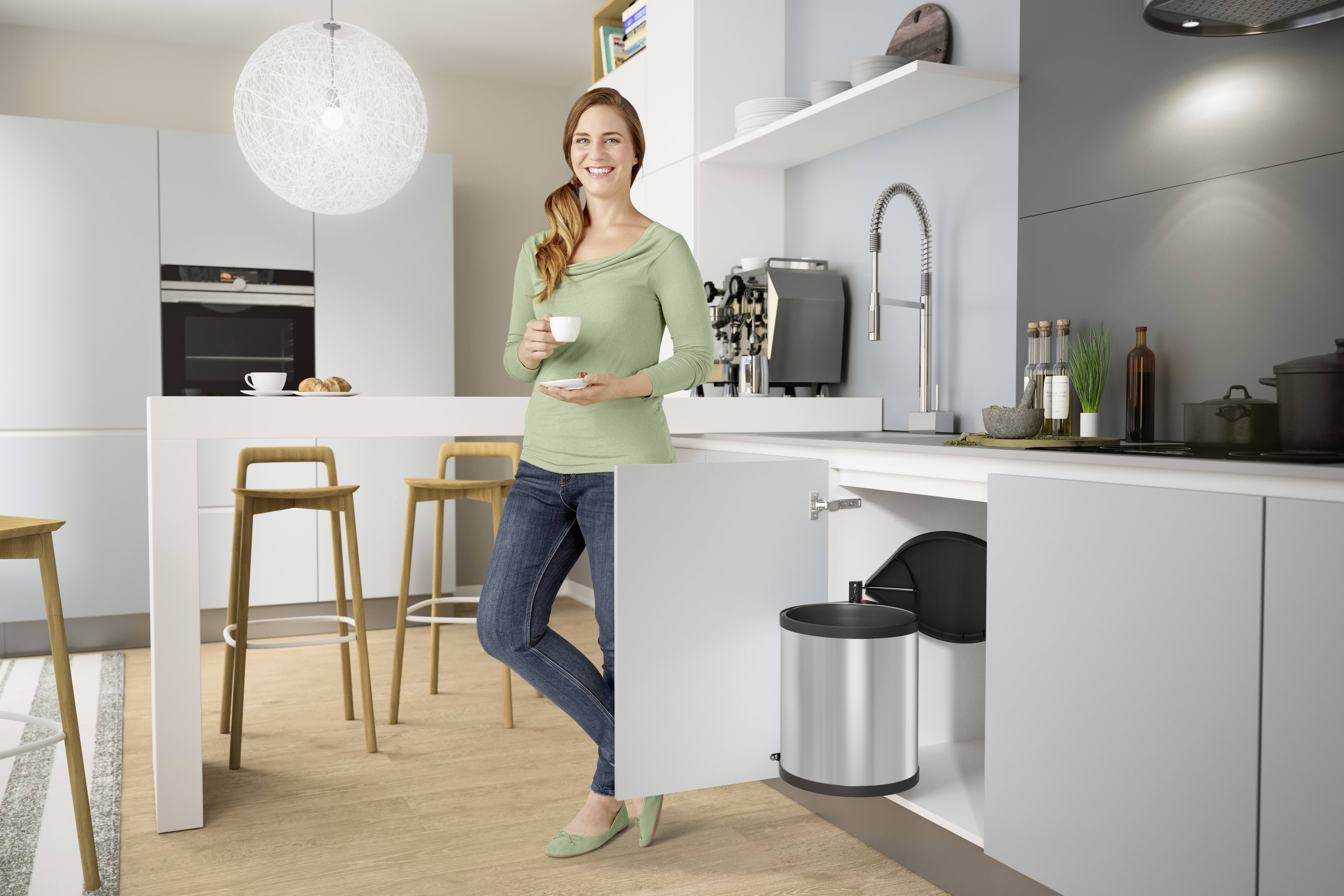 Eine lächelnde Frau steht in einer modernen Küche, hält eine Tasse in der Hand, mit offenem Küchenschrank, der einen Mülleimer zeigt.