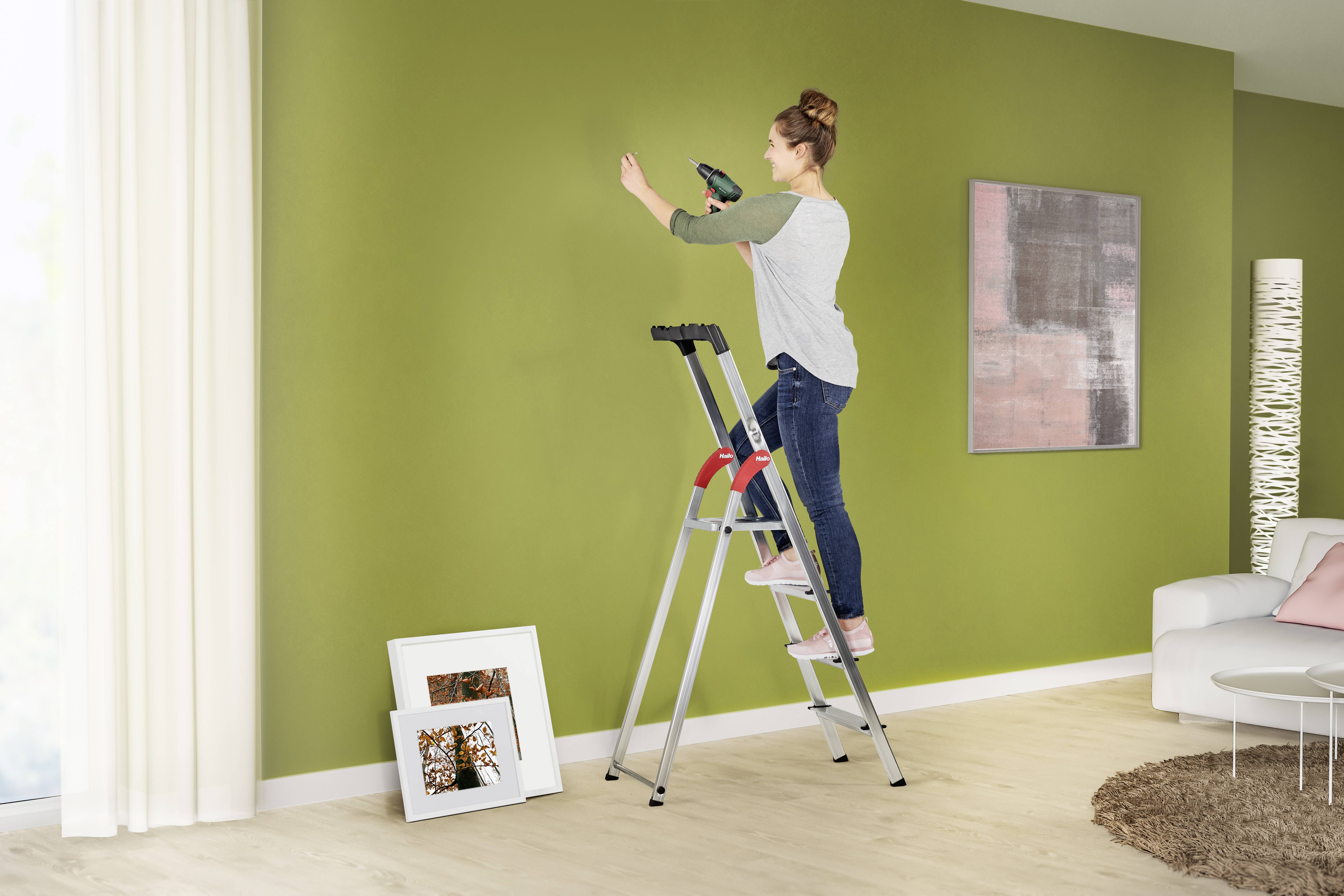 Eine Frau steht auf einer Leiter und bohrt ein Loch in eine grüne Wand. Im Hintergrund stehen gerahmte Bilder und eine Lampe.