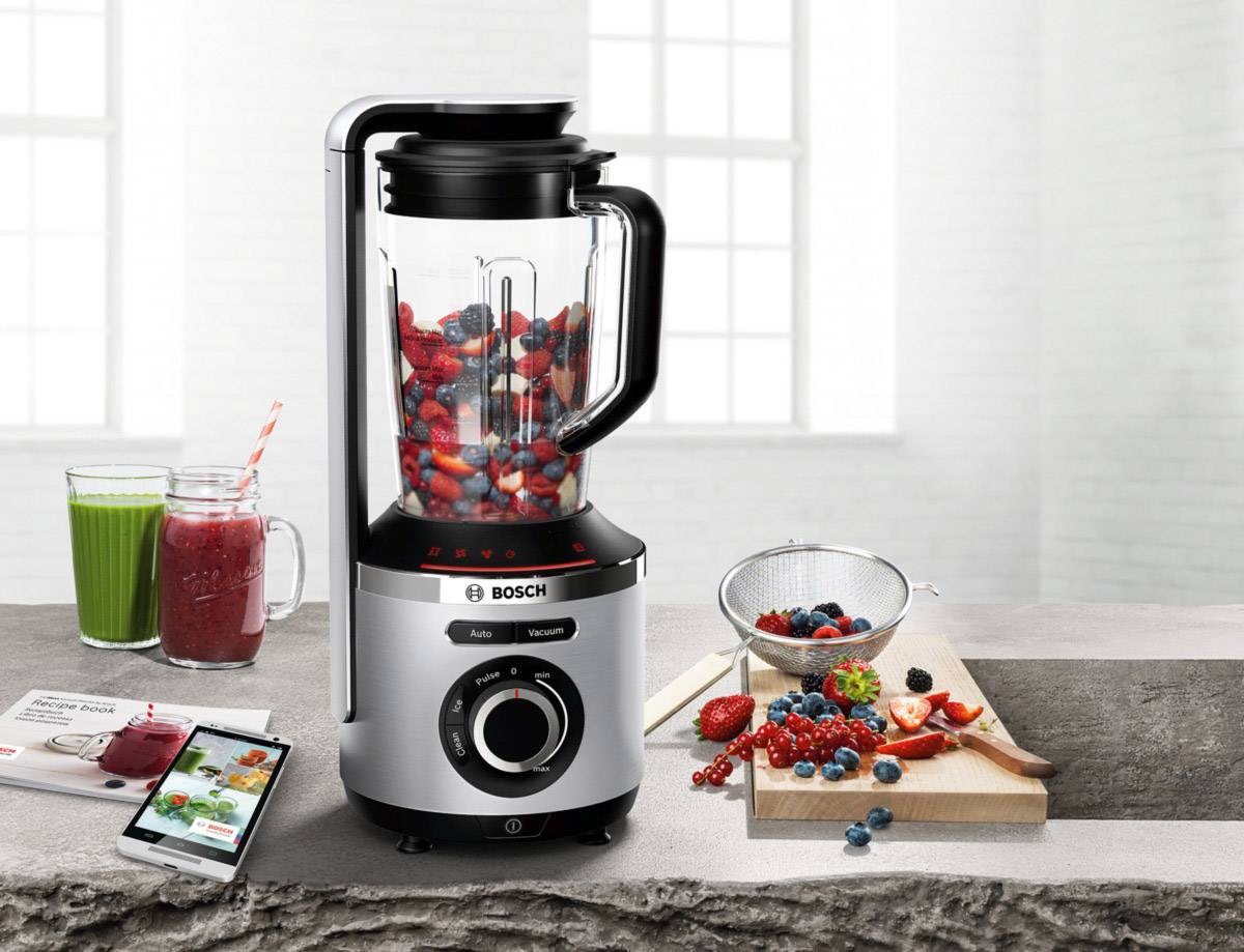 Elektrischer Mixer auf grauem Tisch, gefüllt mit Beeren. Daneben ein Schneidebrett mit Beeren, ein Sieb und zwei Smoothies in Gläsern.