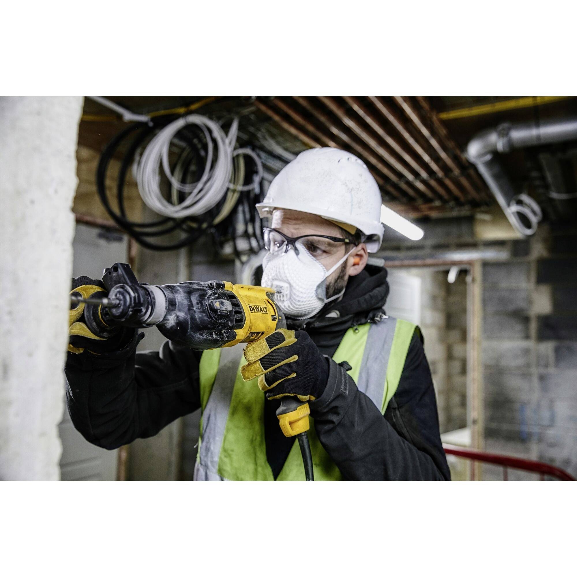 Eine Person in Arbeitskleidung, Schutzmaske und Helm verwendet eine Bohrmaschine in einer Baustelle.