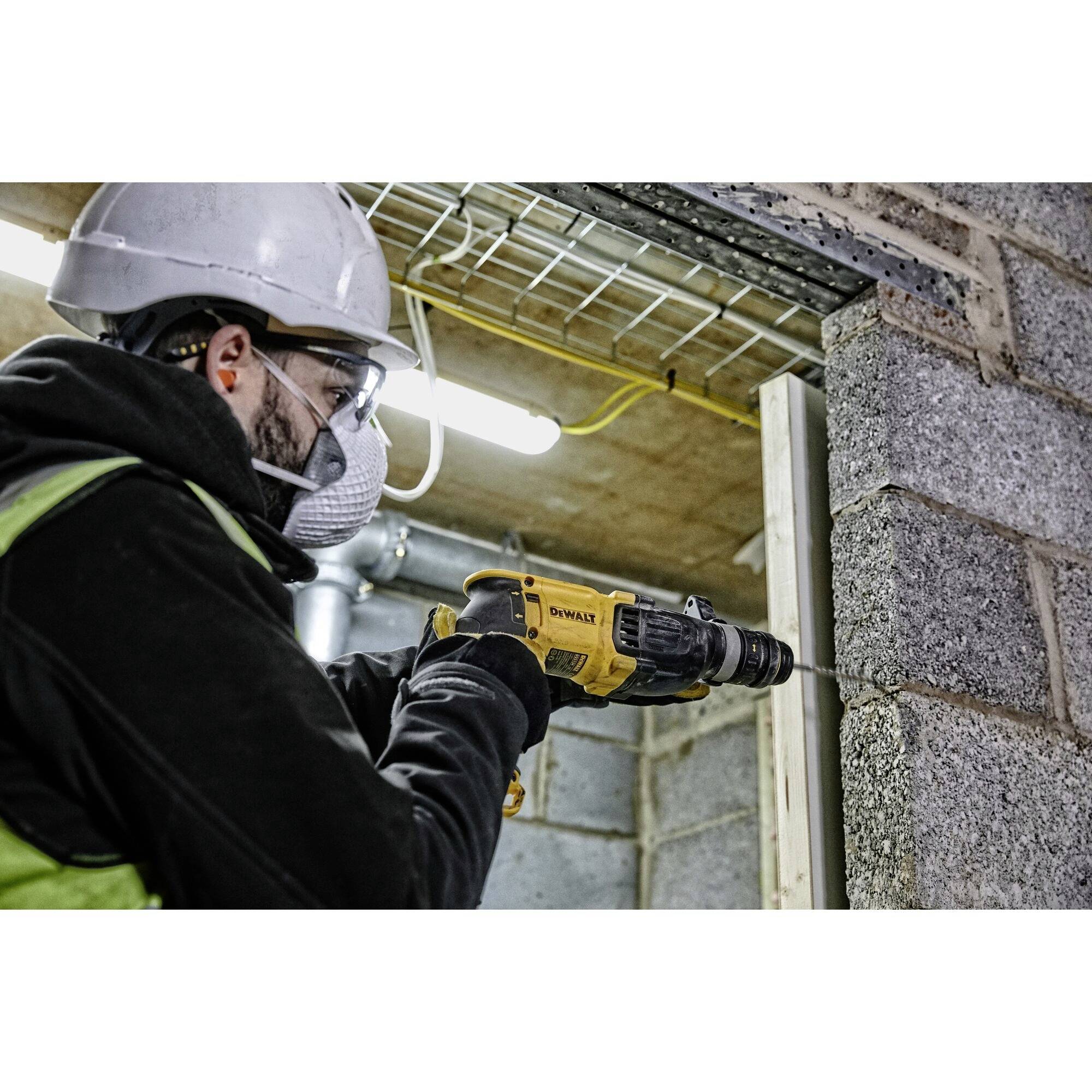 Person im Bauhelm und Schutzbrille bohrt mit einer gelben Bohrmaschine in eine Betonwand. Im Hintergrund sind Kabel sichtbar.