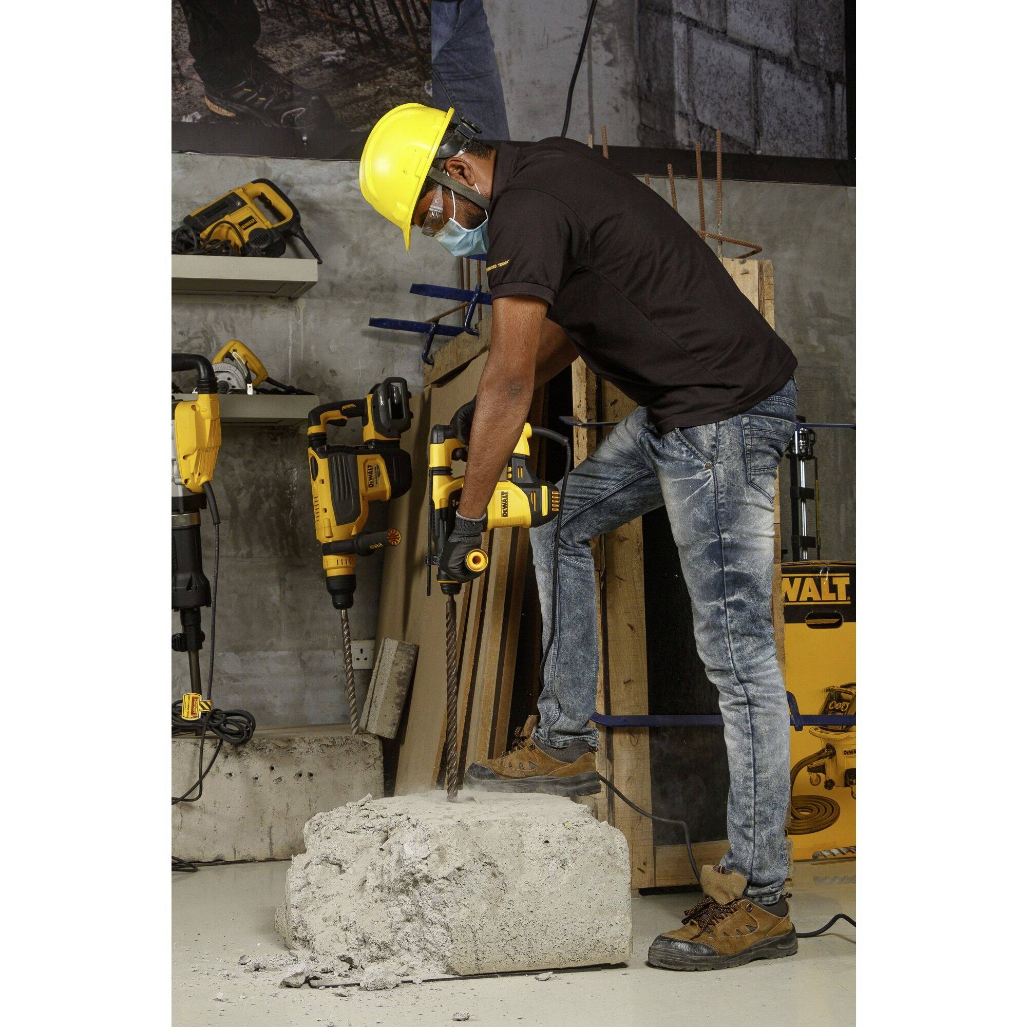 Arbeiter mit Helm und Maske benutzt Bohrmaschine, bearbeitet Betonteil in Werkstattumgebung.