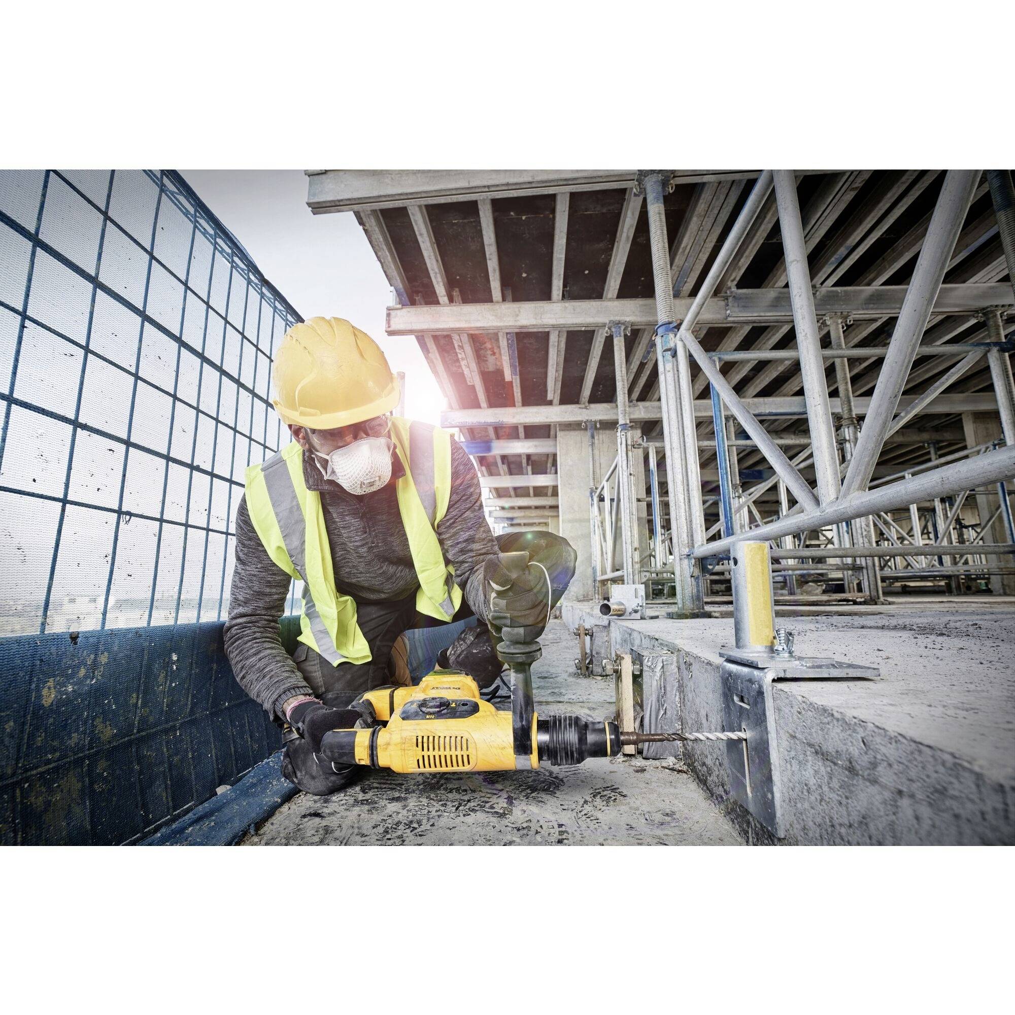 Bauarbeiter trägt Schutzkleidung und verwendet eine Bohrmaschine auf einer Baustelle. Gerüste und unvollendete Strukturen im Hintergrund.