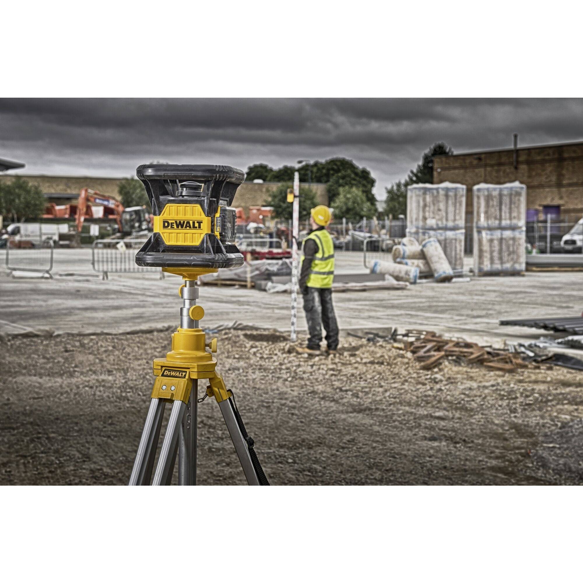 Eine Baustelle mit einem Nivelliergerät auf einem Stativ im Vordergrund. Im Hintergrund misst ein Bauarbeiter mit einer Messlatte.