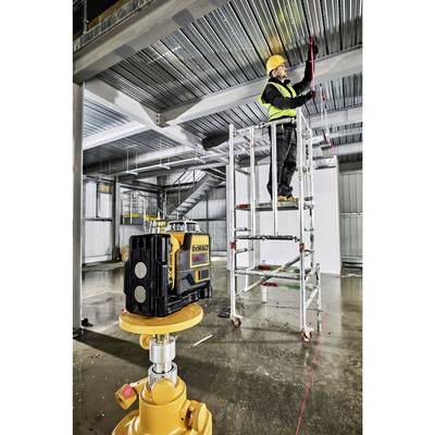 Ein Bauarbeiter steht auf einem Gerüst in einer Halle und misst mit einem Lasergerät die Position einer Metallstruktur an der Decke.