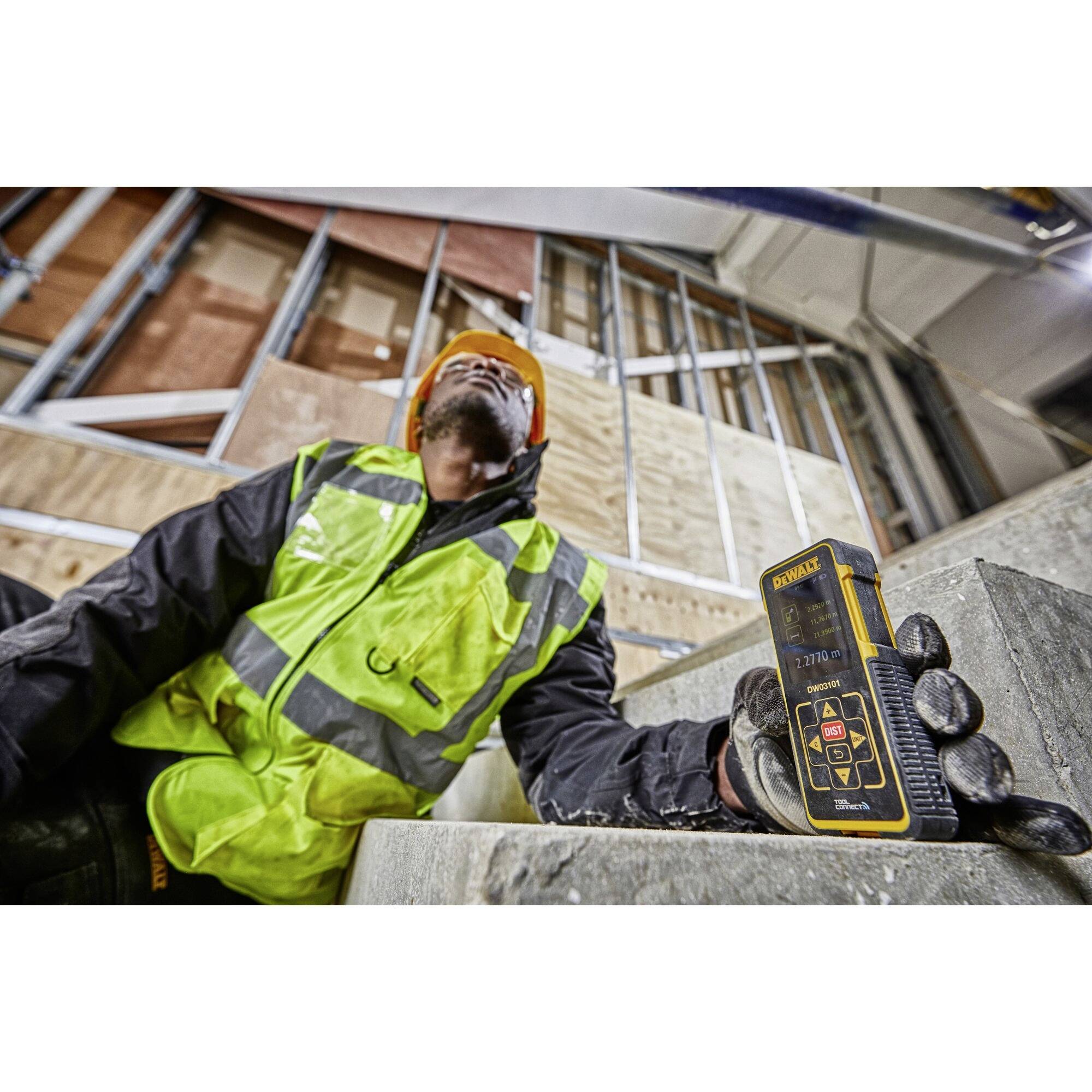 Ein Bauarbeiter mit Helm und Warnweste misst mit einem digitalen Gerät auf einer Baustelle die Entfernung eines Gebäudeteils.