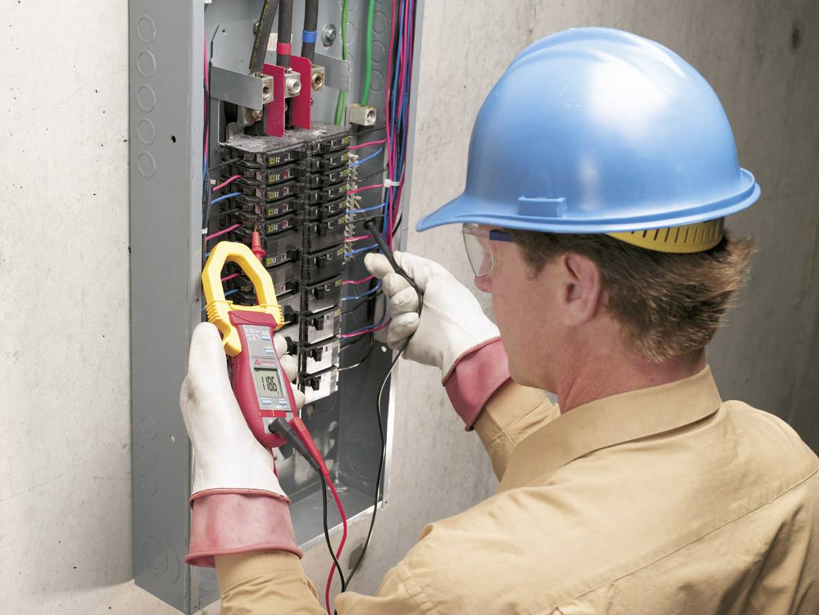 Ein Elektriker mit Schutzhelm und Handschuhen misst Spannung in einem Schaltschrank mit einem digitalen Multimeter.