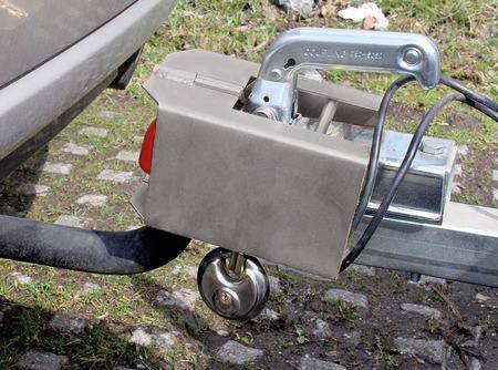 Nahaufnahme einer Anhängerkupplung an einem Auto, die mit einem Anhänger verbunden ist. Gras und Kopfsteinpflaster sind im Hintergrund sichtbar.