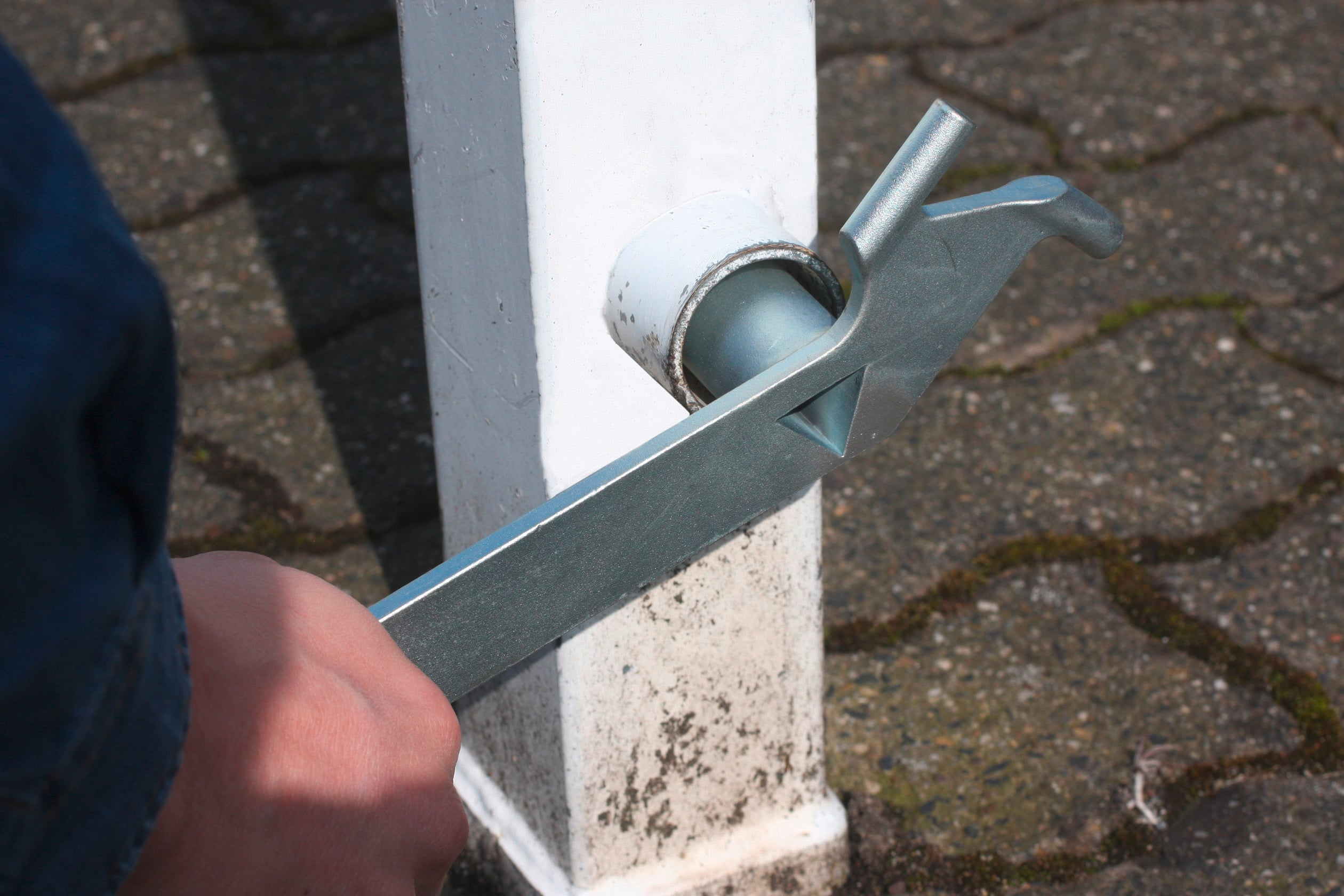 Person zieht mit einem großen Schraubenschlüssel einen Bolzen an einem Metallpfosten fest. Im Hintergrund sind gepflasterte Steine zu sehen.