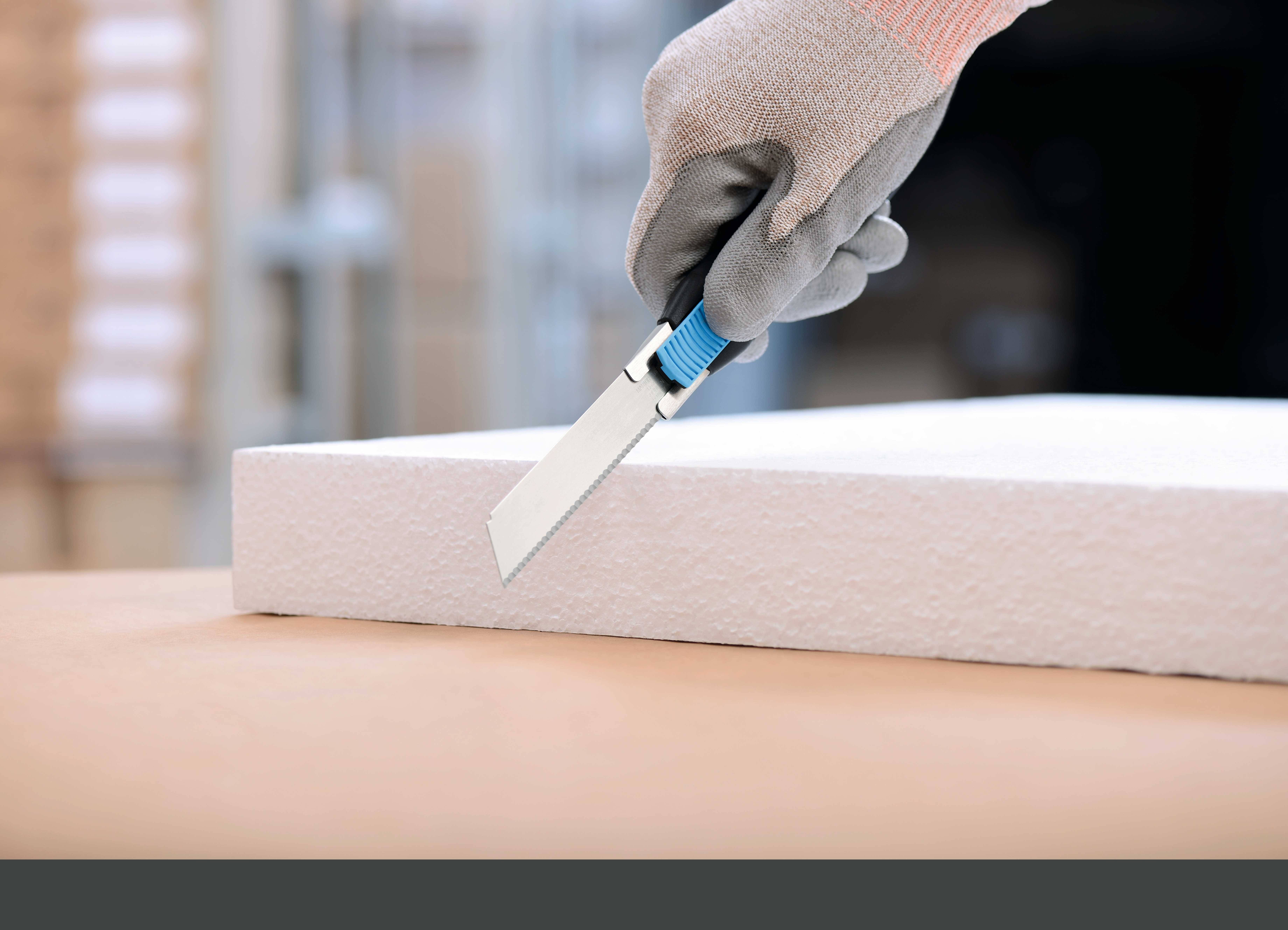 Eine Person schneidet eine Schaumstoffplatte mit einem Cuttermesser. Der Fokus liegt auf der Hand mit Handschuh und dem Messer.