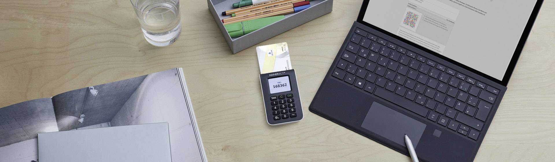 Ein Laptop auf einem Tisch zeigt eine geöffnete Webseite. Daneben liegt ein Kartenterminal mit eingelegter Karte, ein Notizbuch und ein Glas Wasser.