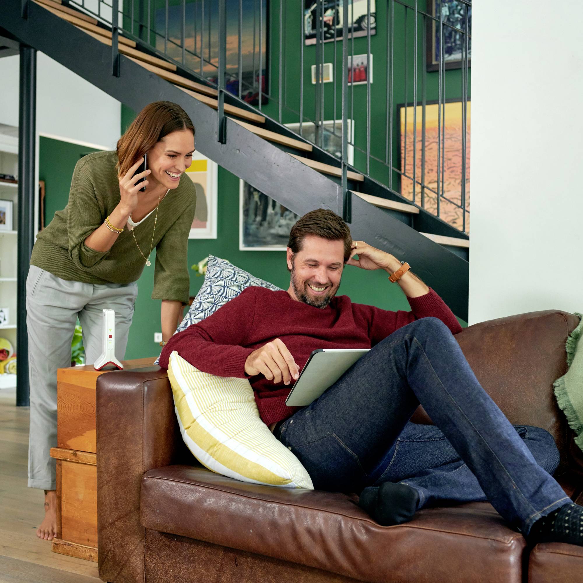 Eine lächelnde Frau telefoniert, während ein Mann auf dem Sofa sitzt, auf einem Tablet lächelt und in einem gemütlichen Wohnzimmer entspannt.