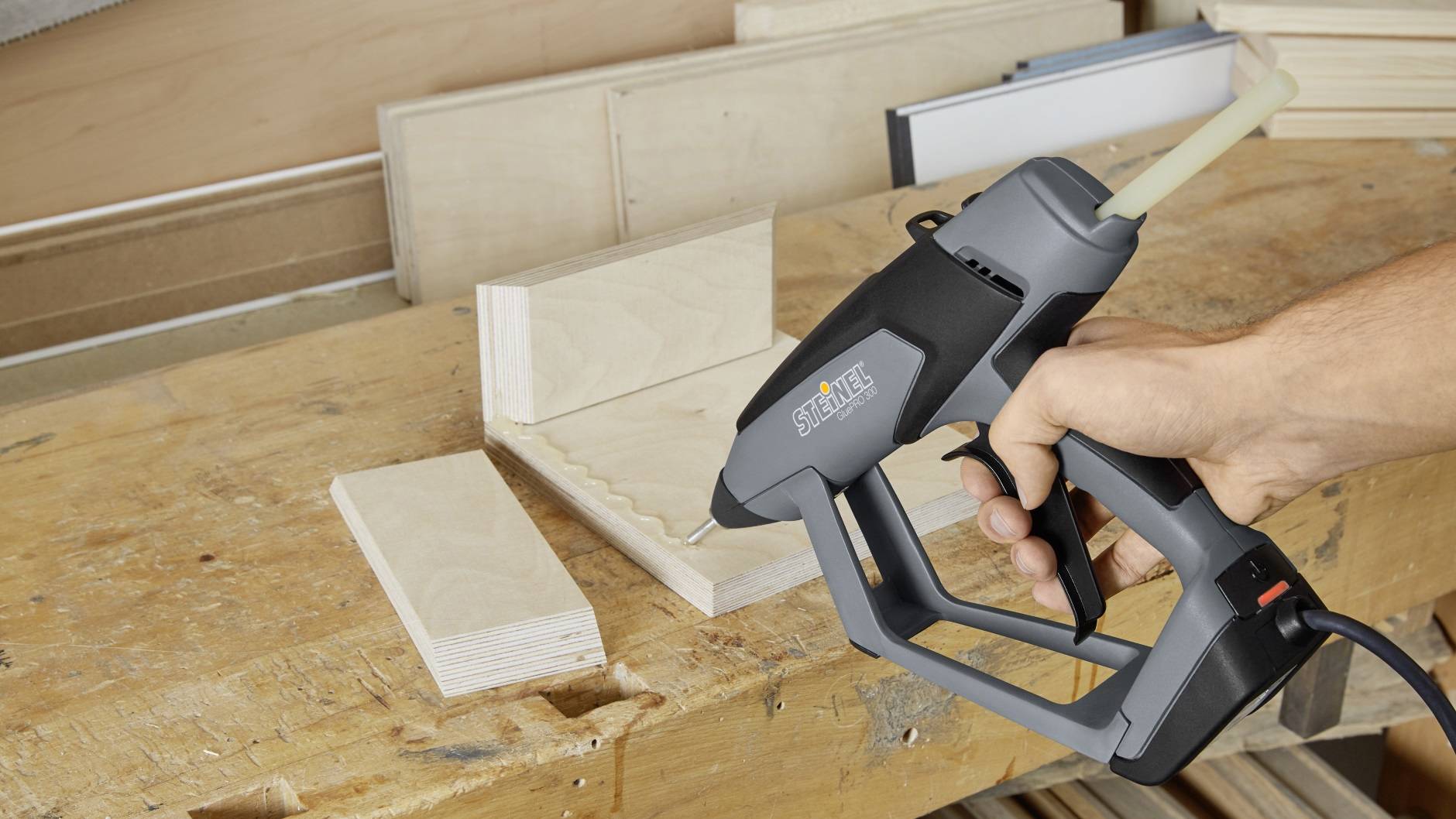 Eine Hand hält eine Heißklebepistole, die auf zwei Holzbretter gerichtet ist, die in einem Winkel zur Holzbearbeitung positioniert sind.
