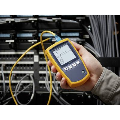 Eine Hand hält ein Netzwerkkabeltester vor einem Server-Rack. Der Tester zeigt Messwerte auf dem Display an. Kabel sind angeschlossen.