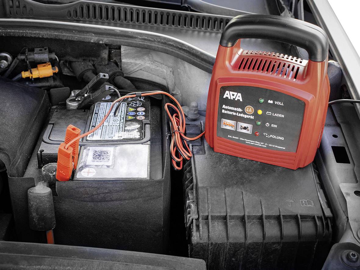 Auto-Motorraum mit Autobatterie und angeschlossenem roten Batterieladegerät. Ladegerät zeigt Ladezustand der Batterie an.