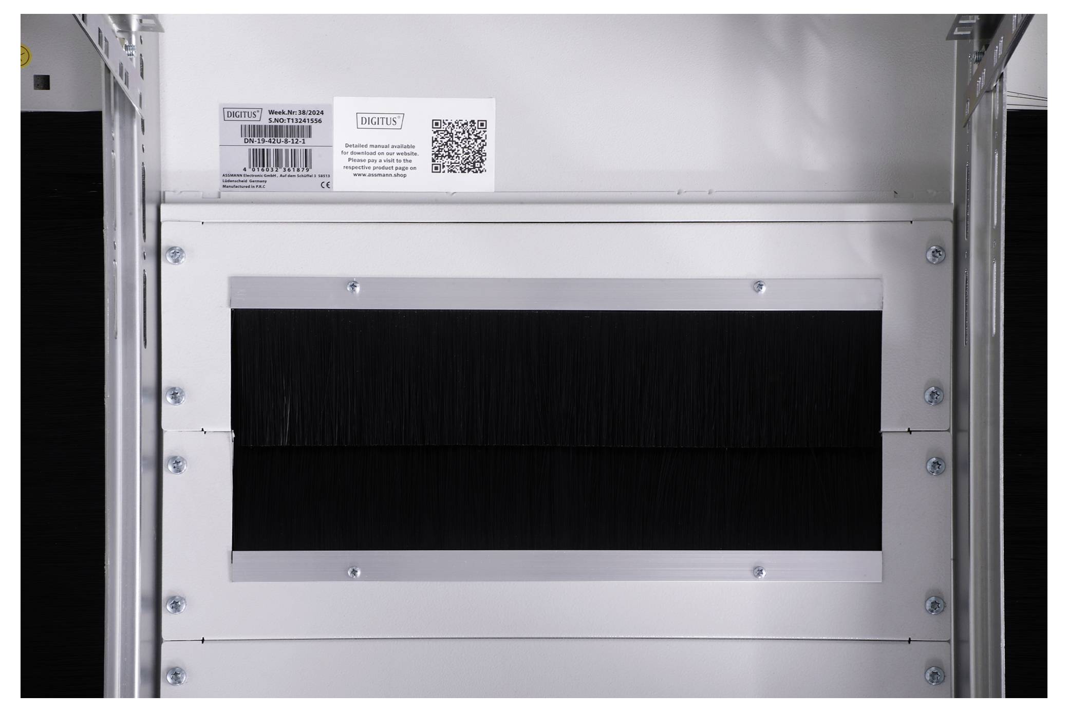Innenansicht eines grauen metallischen Gehäuses mit schwarzem Bürsteneinsatz in der Mitte. Oben ein Etikett mit QR-Code und Produktinformationen.