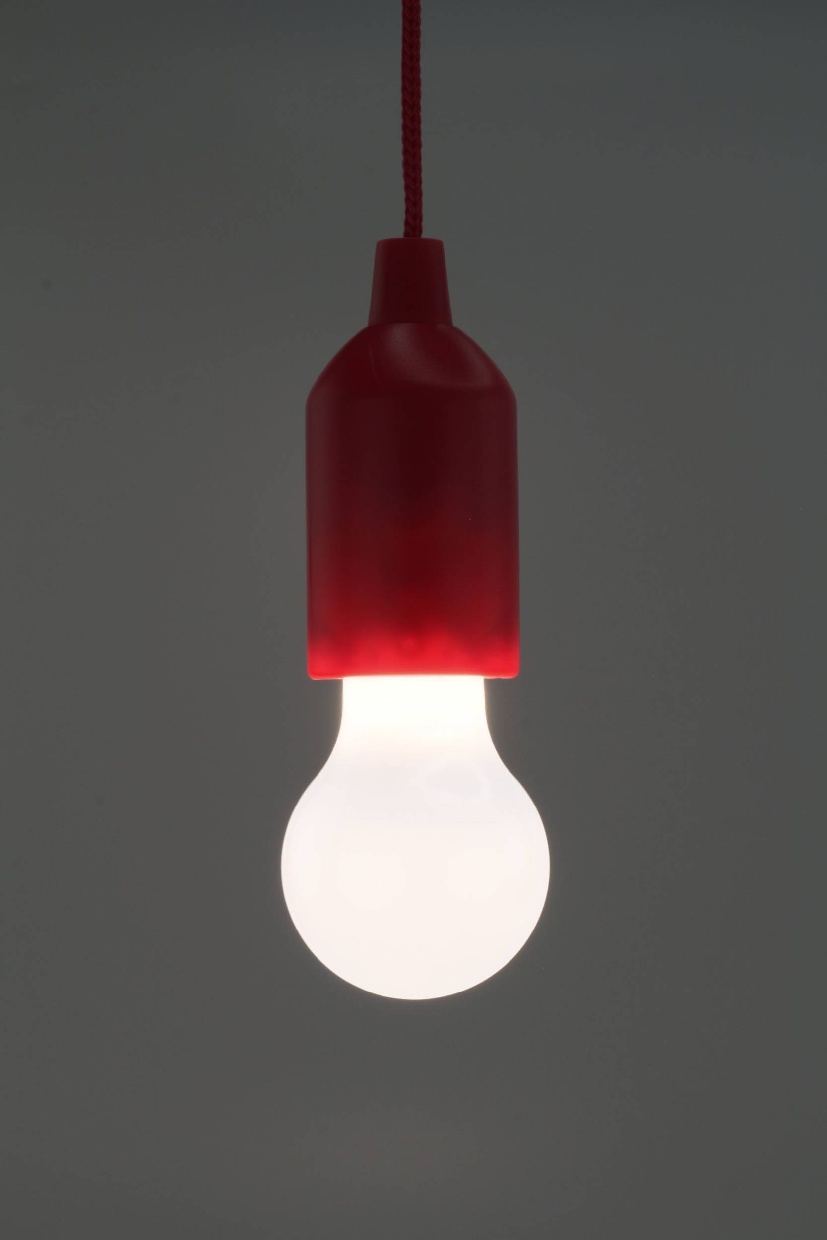 Eine leuchtende Glühbirne mit roter Fassung hängt von der Decke. Hintergrund ist dunkel, Fokus liegt auf der Glühbirne als Lichtquelle.