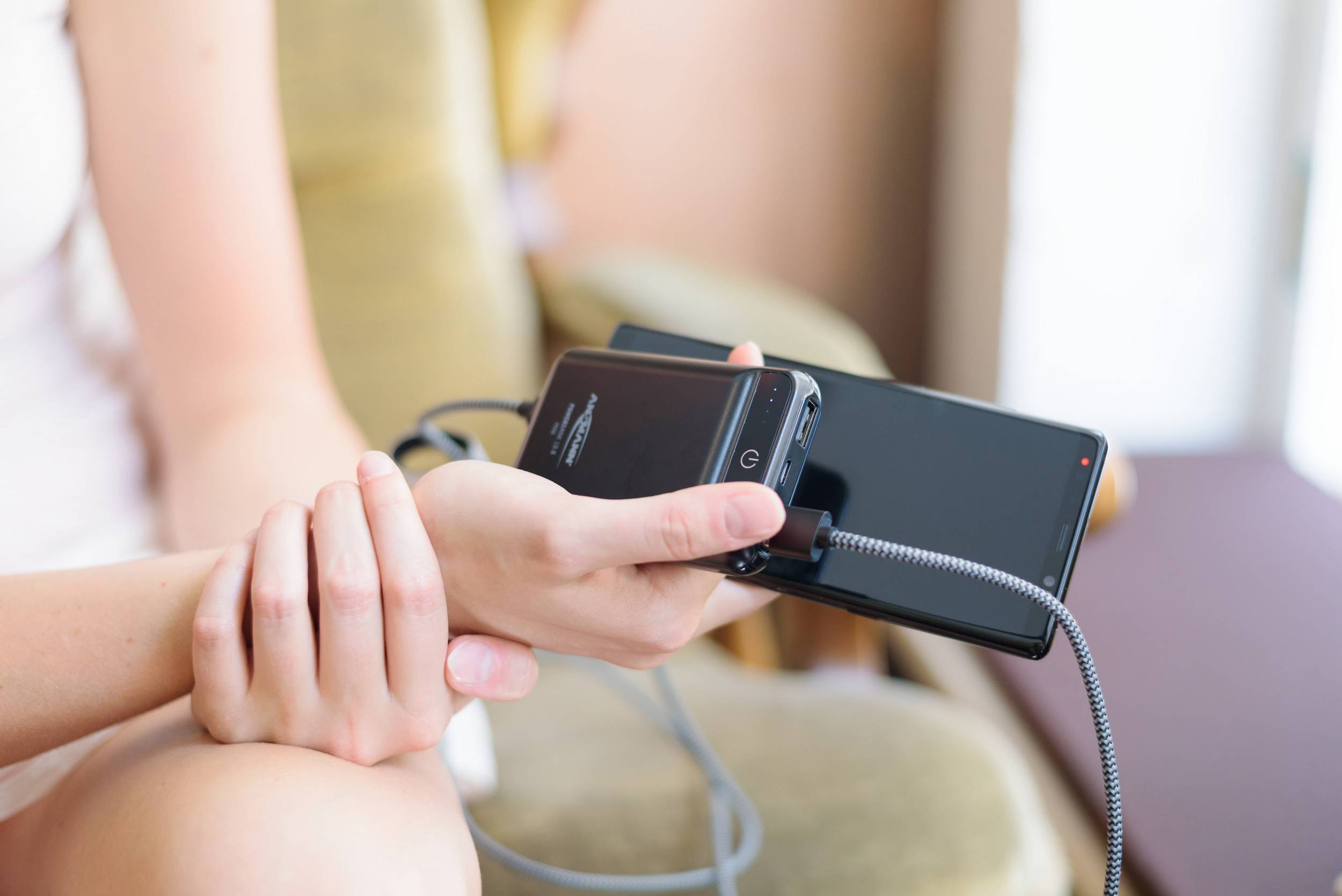 Eine Person hält ein Smartphone, das über ein Kabel mit einem tragbaren Ladegerät verbunden ist, während sie auf einem Stuhl sitzt.