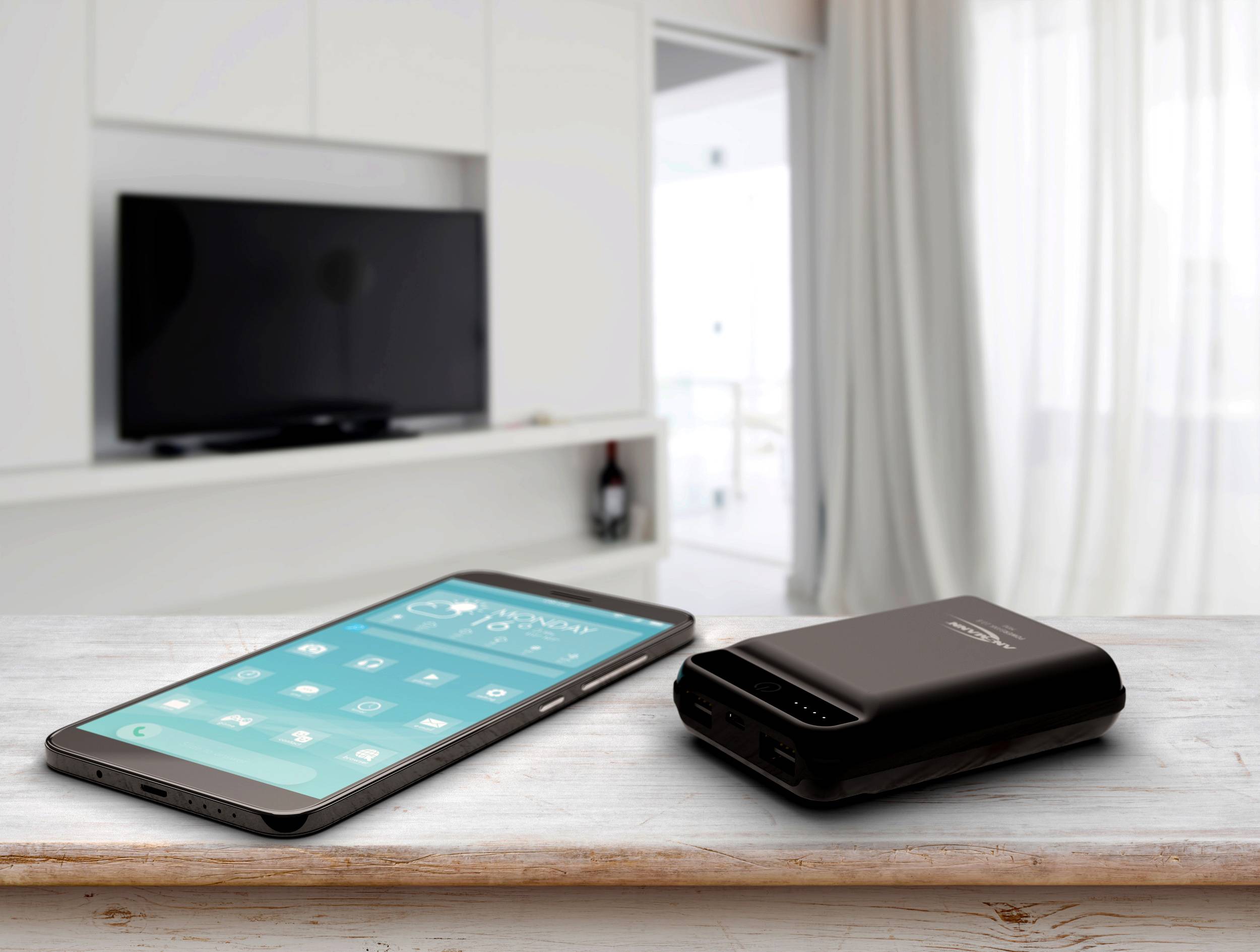 Ein Smartphone und eine Powerbank liegen auf einem Holztisch vor einem modernen Wohnzimmer mit einem Fernseher im Hintergrund.