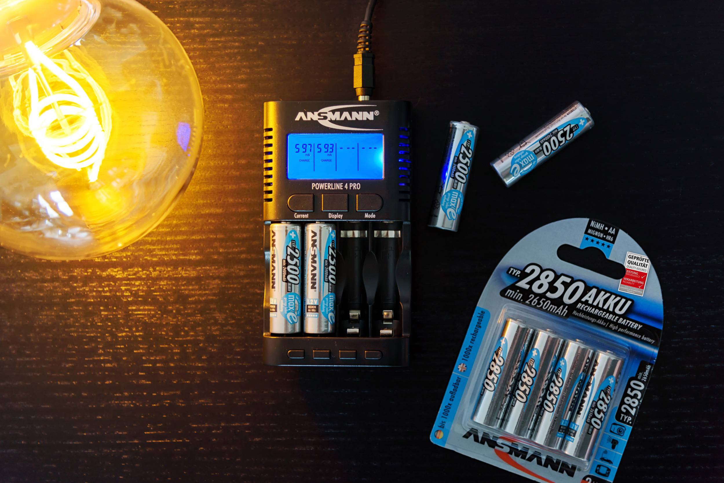 Akku-Ladegerät lädt vier AA-Akkus auf schwarzem Tisch neben einer Schachtel mit zusätzlichen Batterien und einer leuchtenden Glühbirne.