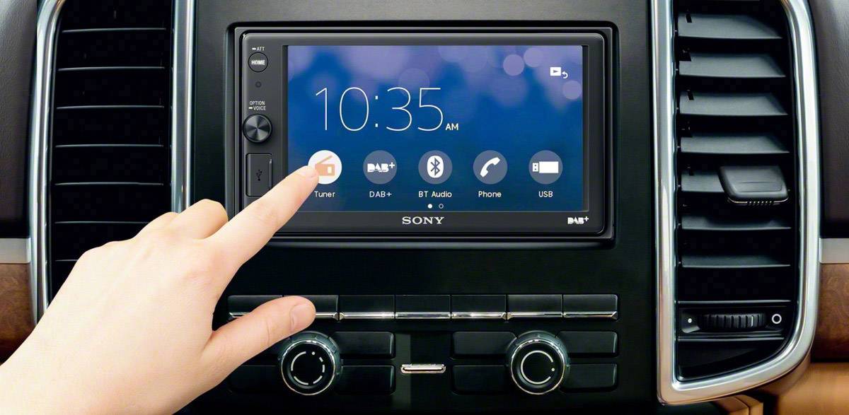 Person tippt auf Touchscreen in einem Auto. Display zeigt Uhrzeit '10:35 AM'. Symbole für Tuner und DAB+ sind sichtbar.
