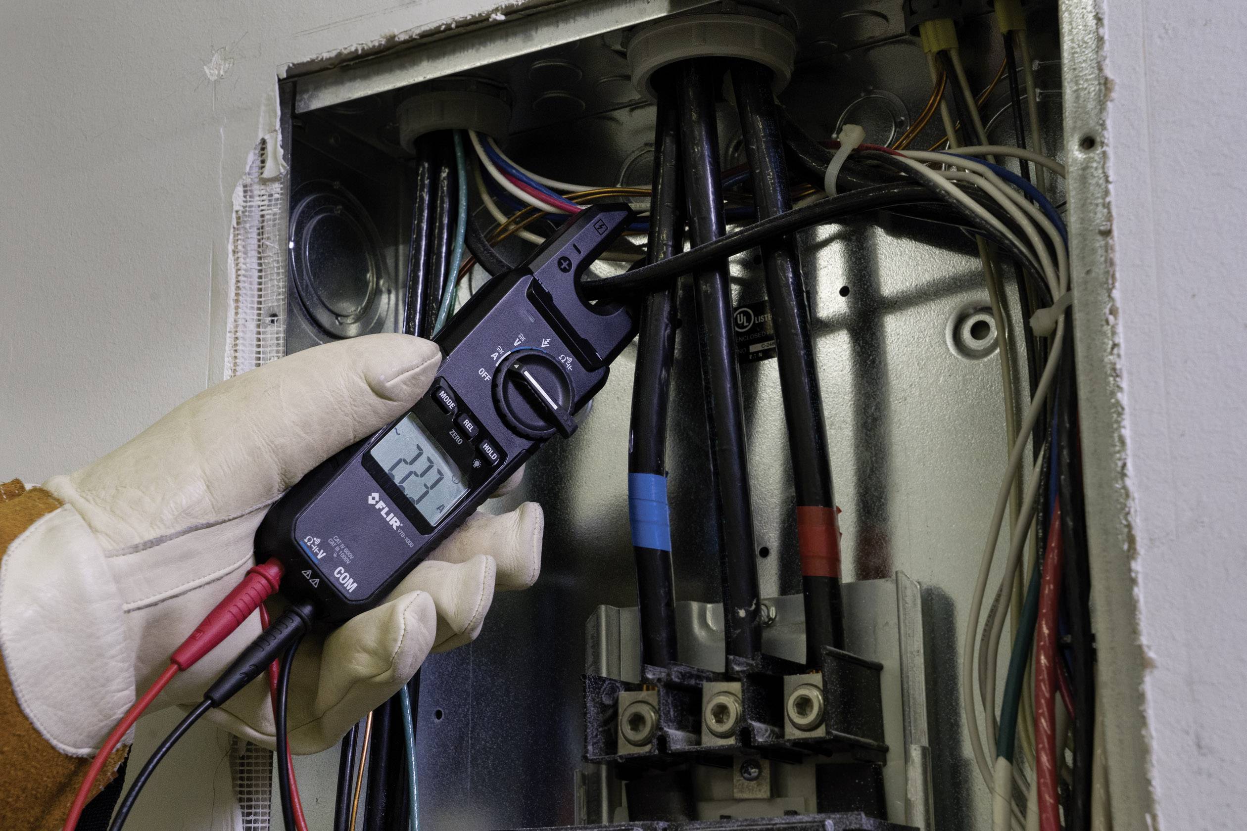 Eine Person überprüft die Spannung eines Sicherungskastens mit einem Multimeter; das Display zeigt 222 Volt an.