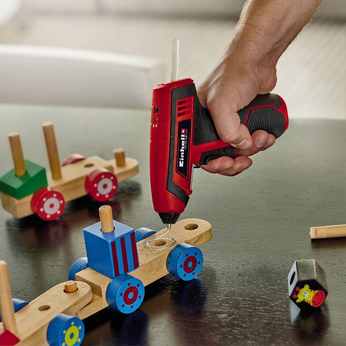 Eine Person verwendet eine Heißklebepistole, um ein Holzspielzeug auf einem Tisch zu reparieren. Mehrere Holzspielzeuge sind im Hintergrund sichtbar.