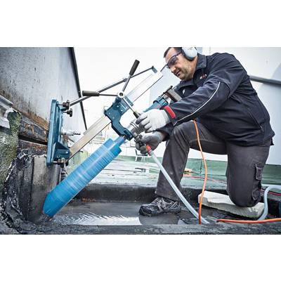 Eine Person arbeitet mit einer Bohrmaschine auf einem Flachdach, trägt Schutzkleidung und Gehörschutz, während sie in Beton bohrt.