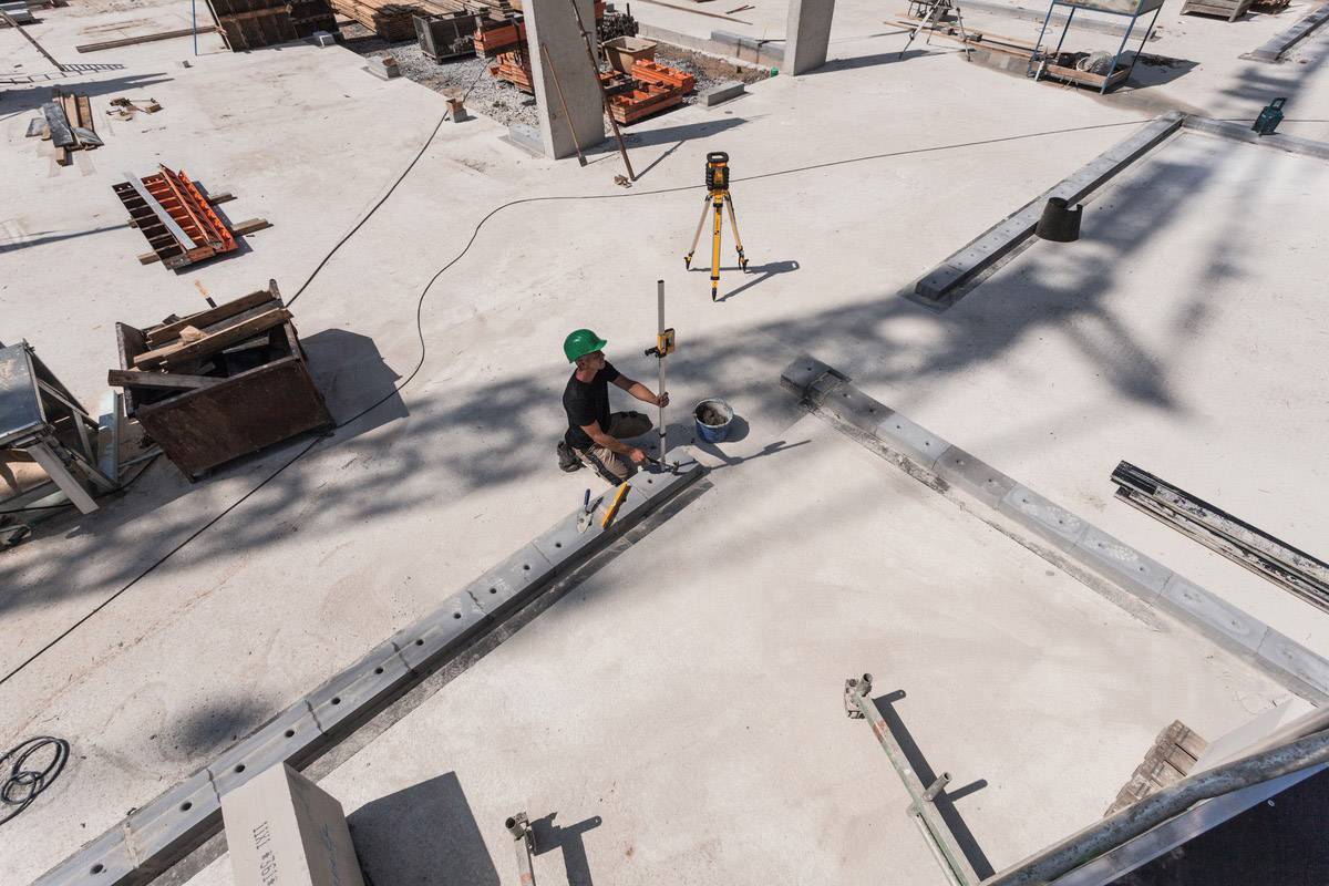 Eine Person mit grünem Helm misst auf einer Baustelle die Höhe mit einem Nivelliergerät. Umgebende Materialien und Werkzeuge sind sichtbar.
