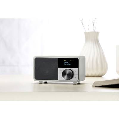Ein weißes Digitalradio mit schwarzem Bildschirm zeigt 'Kaffen' und 'Time to Chill'. Es steht auf einem Tisch neben einem Buch und einer Vase.