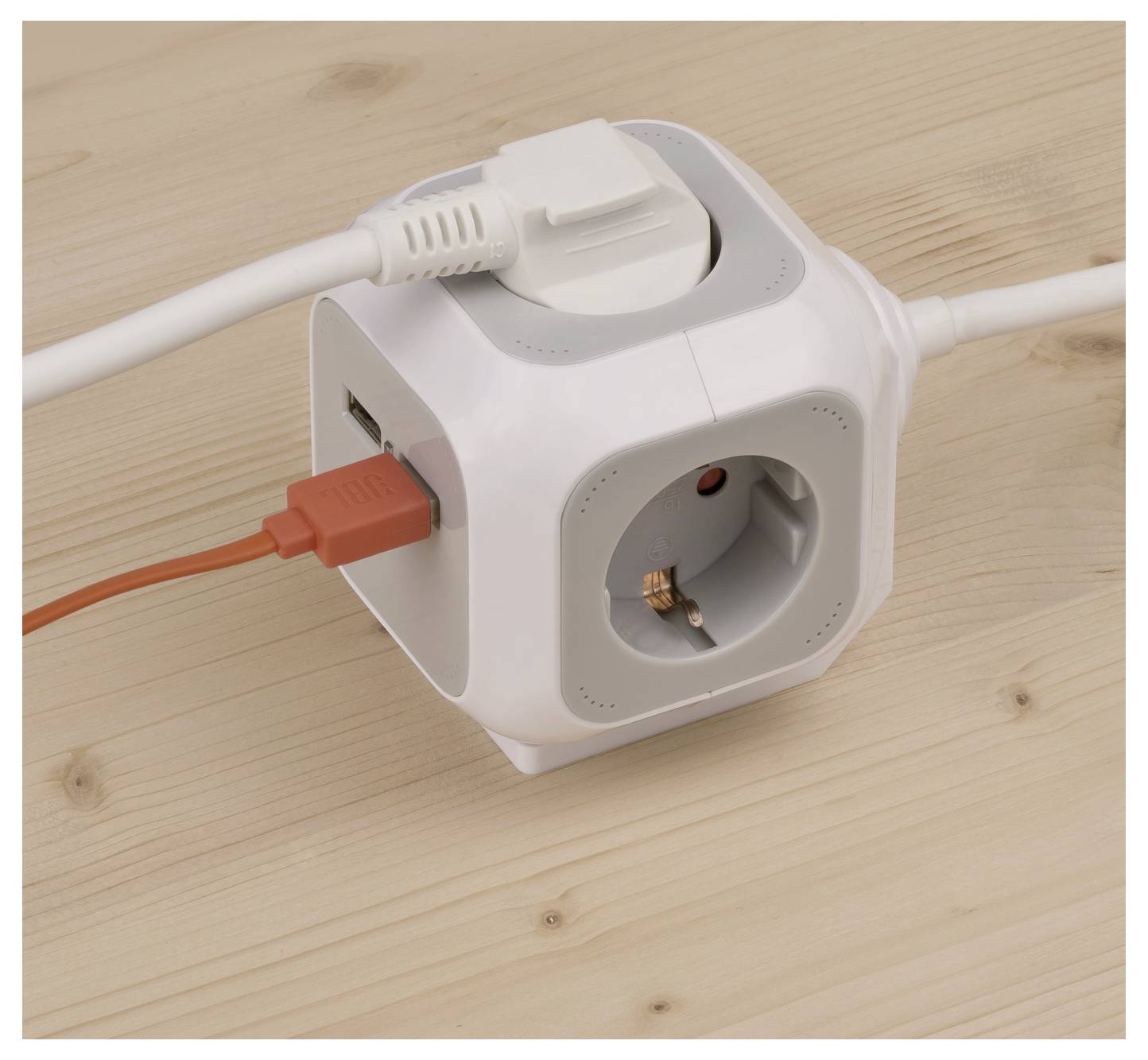 Weiße würfelförmige Steckdosenleiste auf einem Holztisch mit mehreren angeschlossenen Steckern, einschließlich eines USB-Kabels und einer europäischen Steckdose.