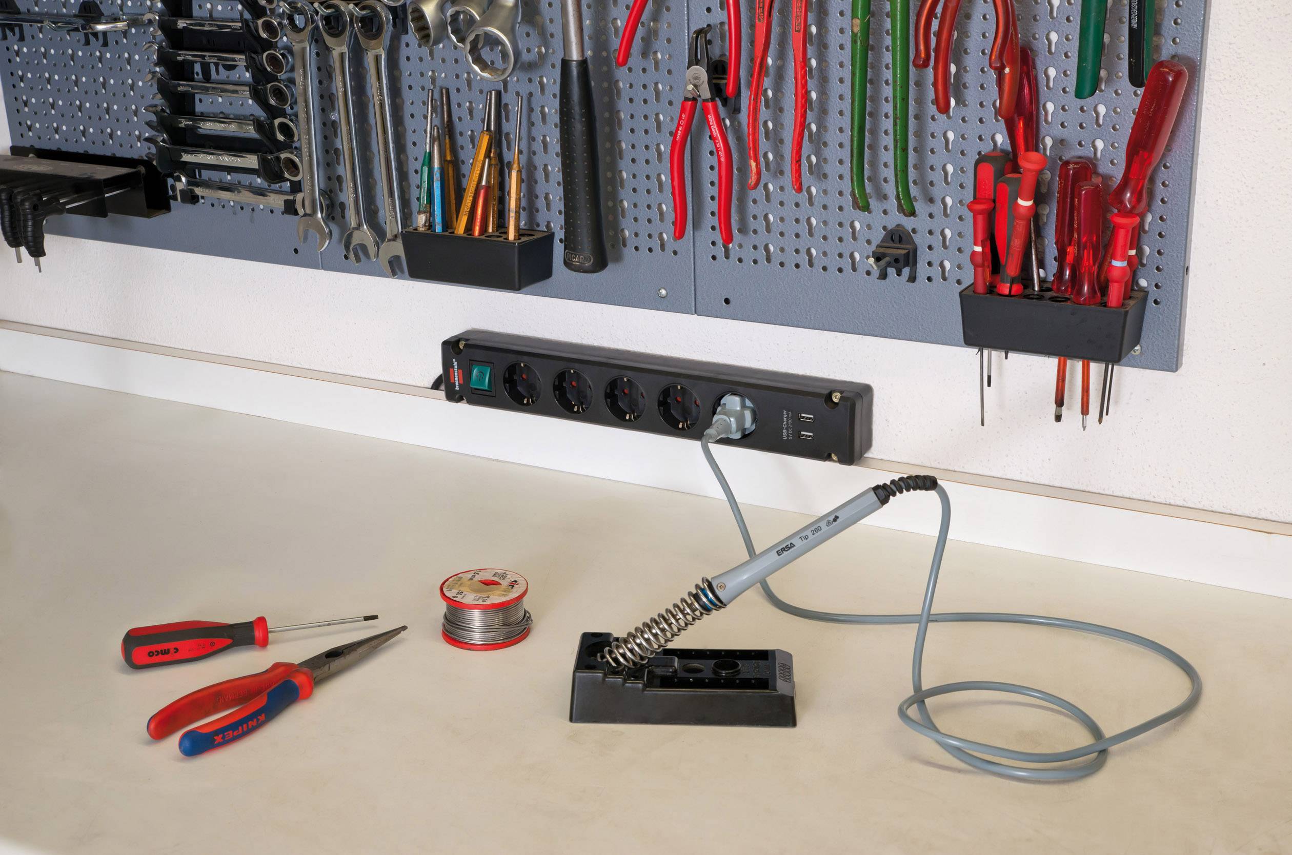 Werkbank mit Lötstation, Lötkolben in Halterung, Schraubendreher und Lötzinn auf der Tischfläche, Werkzeugwand im Hintergrund.
