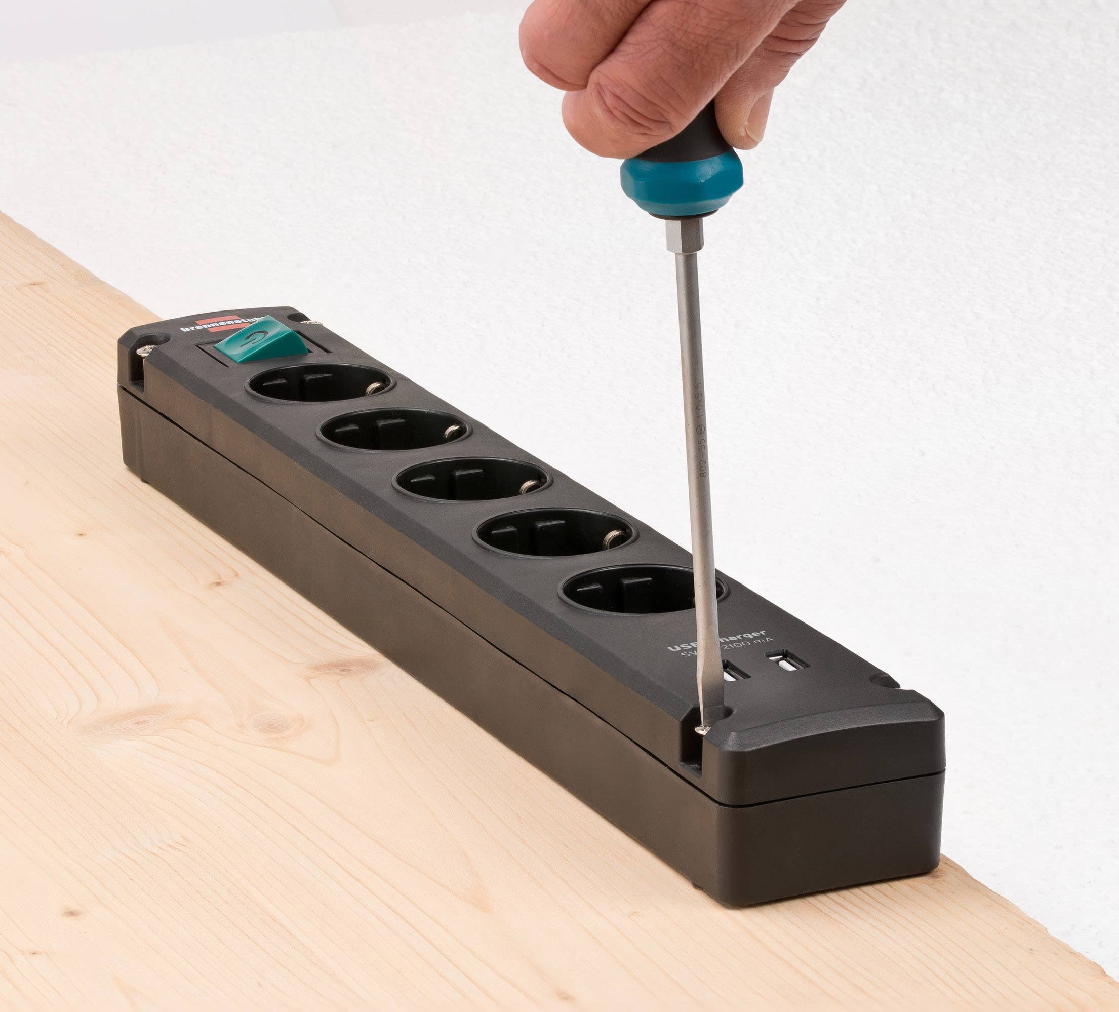 Person dreht mit einem Schraubendreher die Schraube an einer schwarzen Mehrfachsteckdose auf einem Holztisch auf.