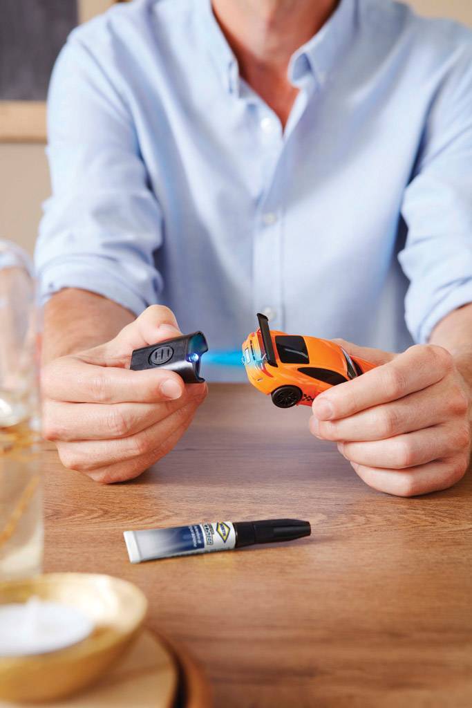 Eine Person klebt ein kleines, orangefarbenes Spielzeugauto mit einem blauen Klebestift auf einem Holztisch.