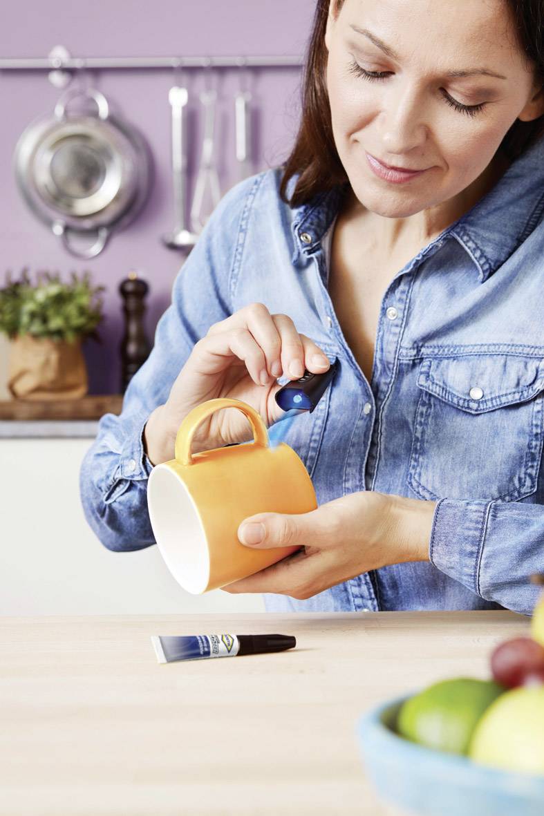 Frau in Jeanshemd repariert orange Tasse mit Kleber in gemütlicher Küche, Werkzeuge und Gewürze im Hintergrund.