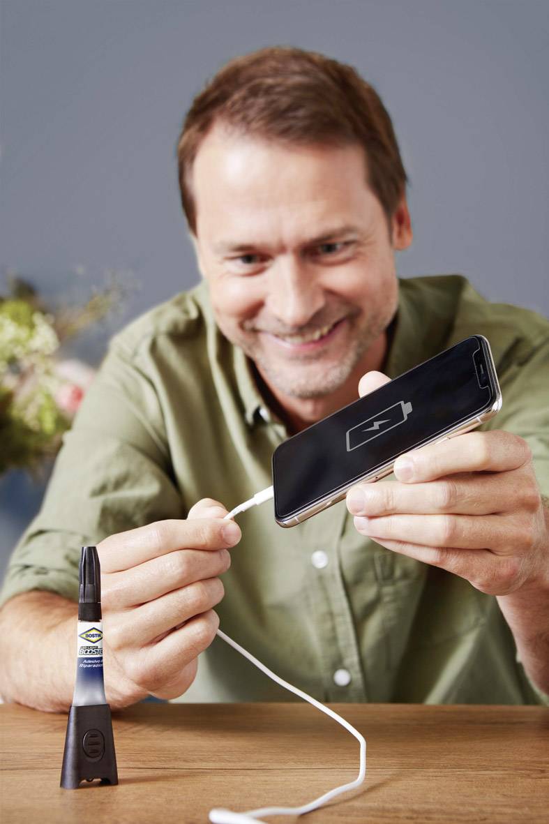 Ein Mann lächelt, während er ein Smartphone auflädt. Das Telefon zeigt ein Ladesymbol. Im Hintergrund ist eine Pflanze sichtbar.