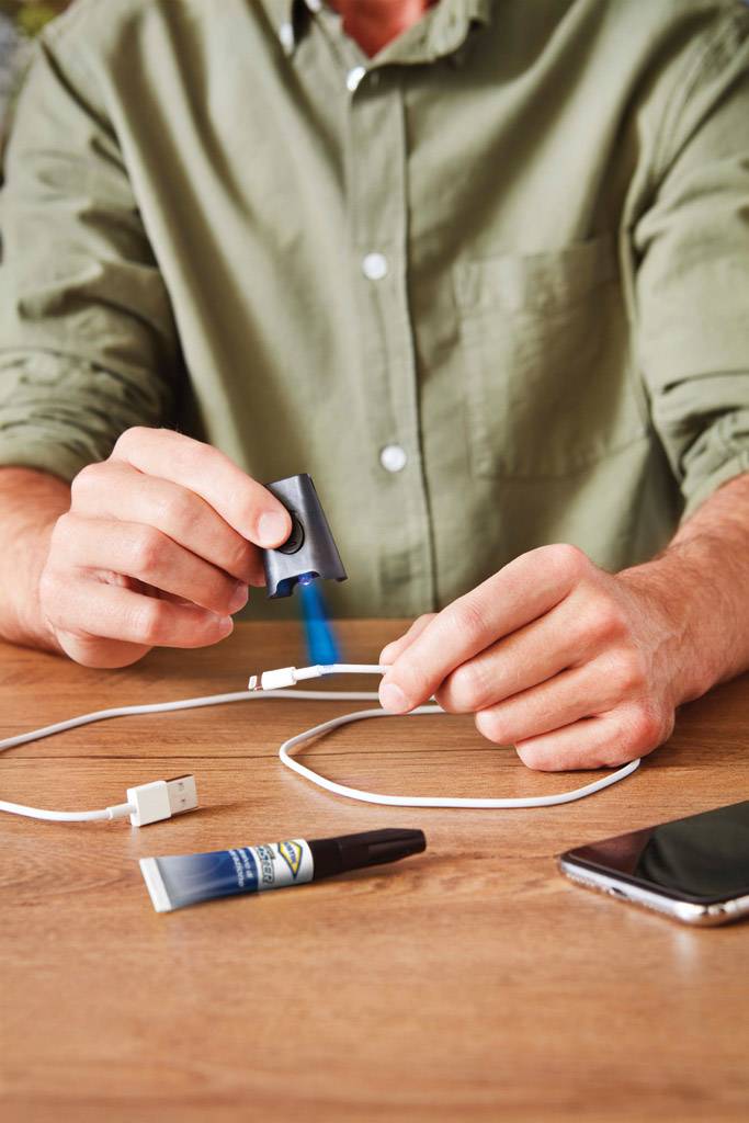 Eine Person steckt einen USB-Stecker in einen Adapter ein, der auf einem Holztisch liegt. Daneben liegen ein Smartphone und ein kleiner Kleber.