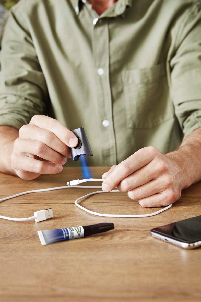 Eine Person repariert ein Ladekabel mit Klebstoff und einem blauen Reparaturstift. Auf dem Tisch liegt ein Smartphone.