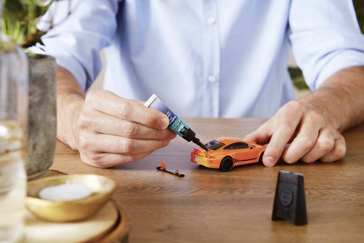 Eine Person repariert ein orangefarbenes Spielzeugauto mit Kleber auf einem Holztisch. Im Hintergrund sind Pflanzen zu sehen.